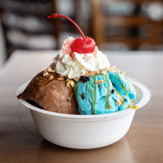 An ice cream sundae with blue and brown ice cream in a paper dish topped with peanuts, caramel, whipped cream, and a cherry. 