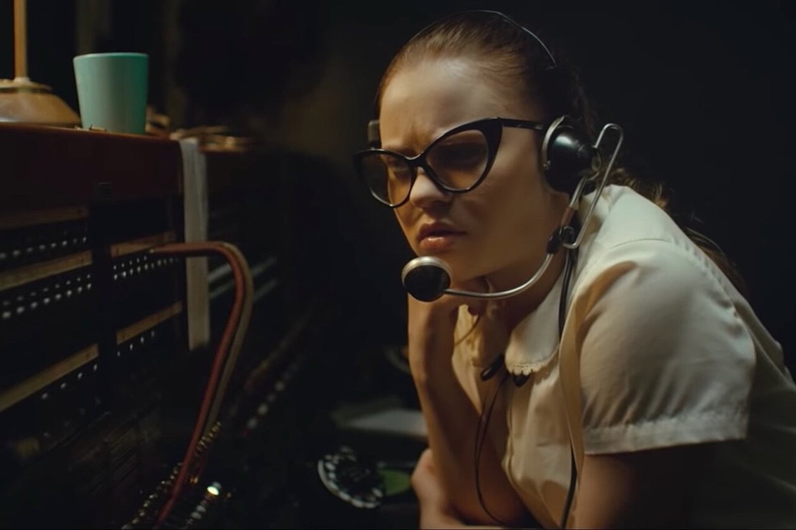 A young woman sits at a old-fashioned telephone switchboard wearing headphones.