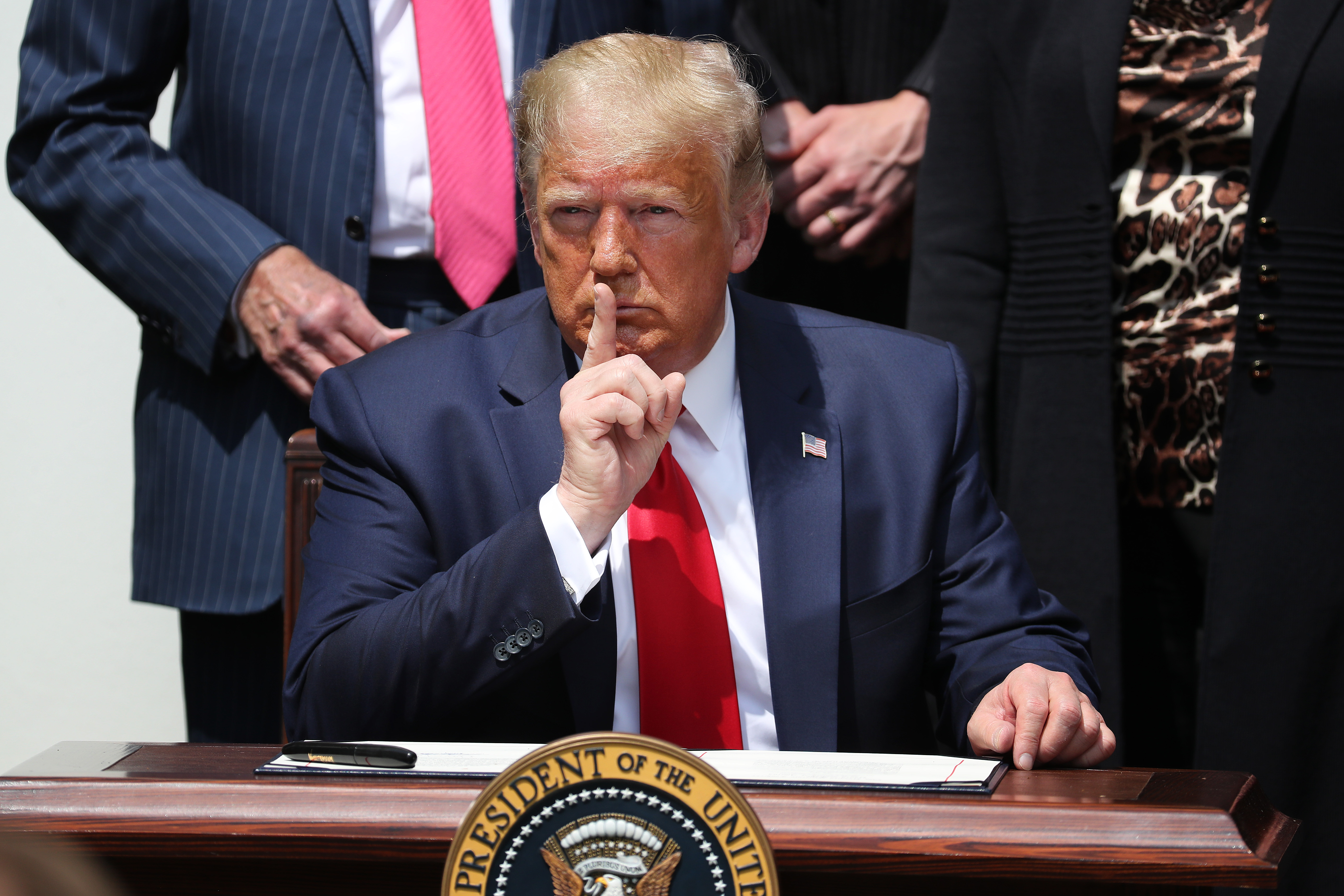 President Trump sitting at a desk holding his finger to his lips in a shushing gesture.