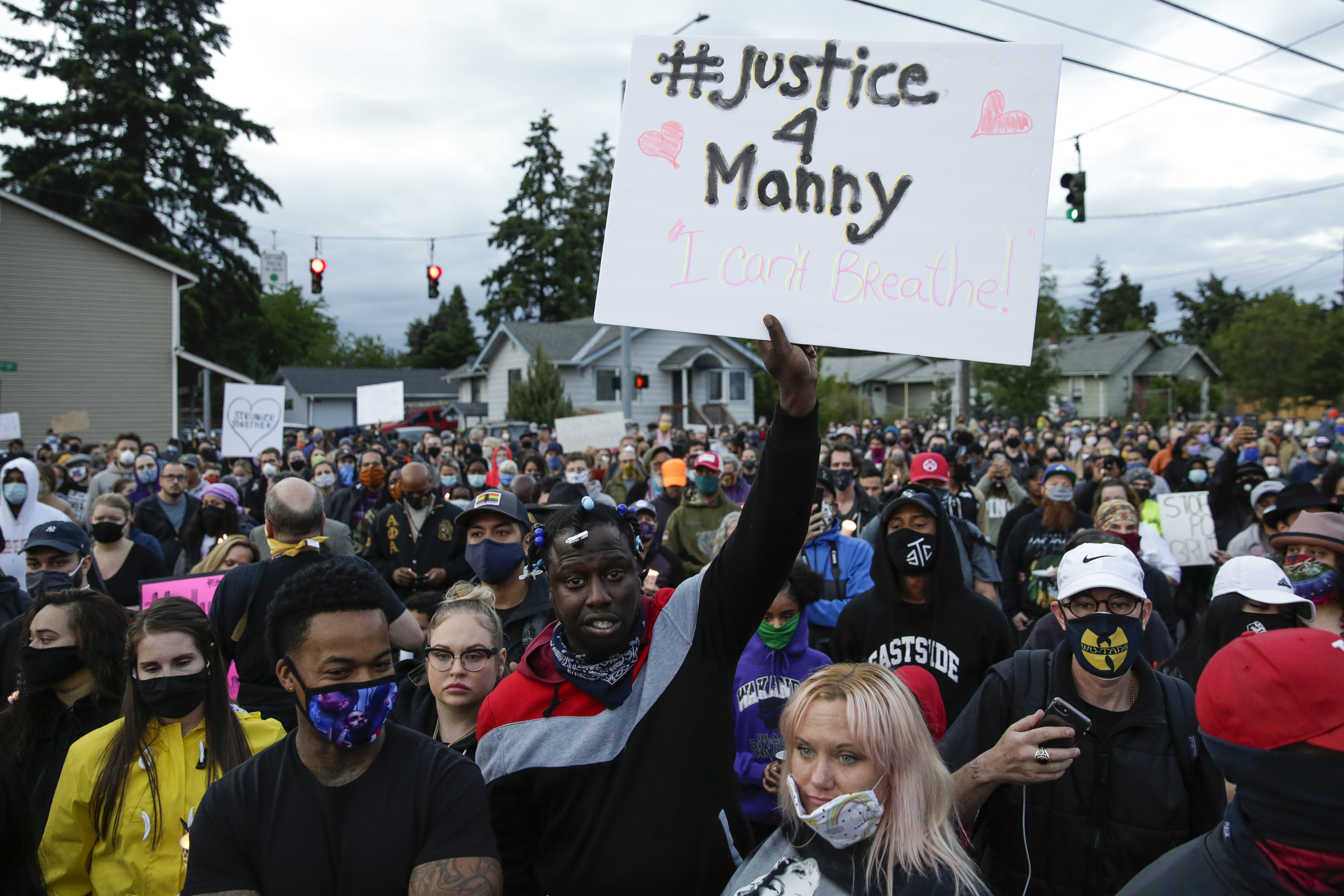 A diverse, masked crowd is packed onto a street under a cloudy sky. A black man with short braids stands at the front of the group, and raises a white sign with black letters and pink hearts reading, “#Justice 4 Manny.”