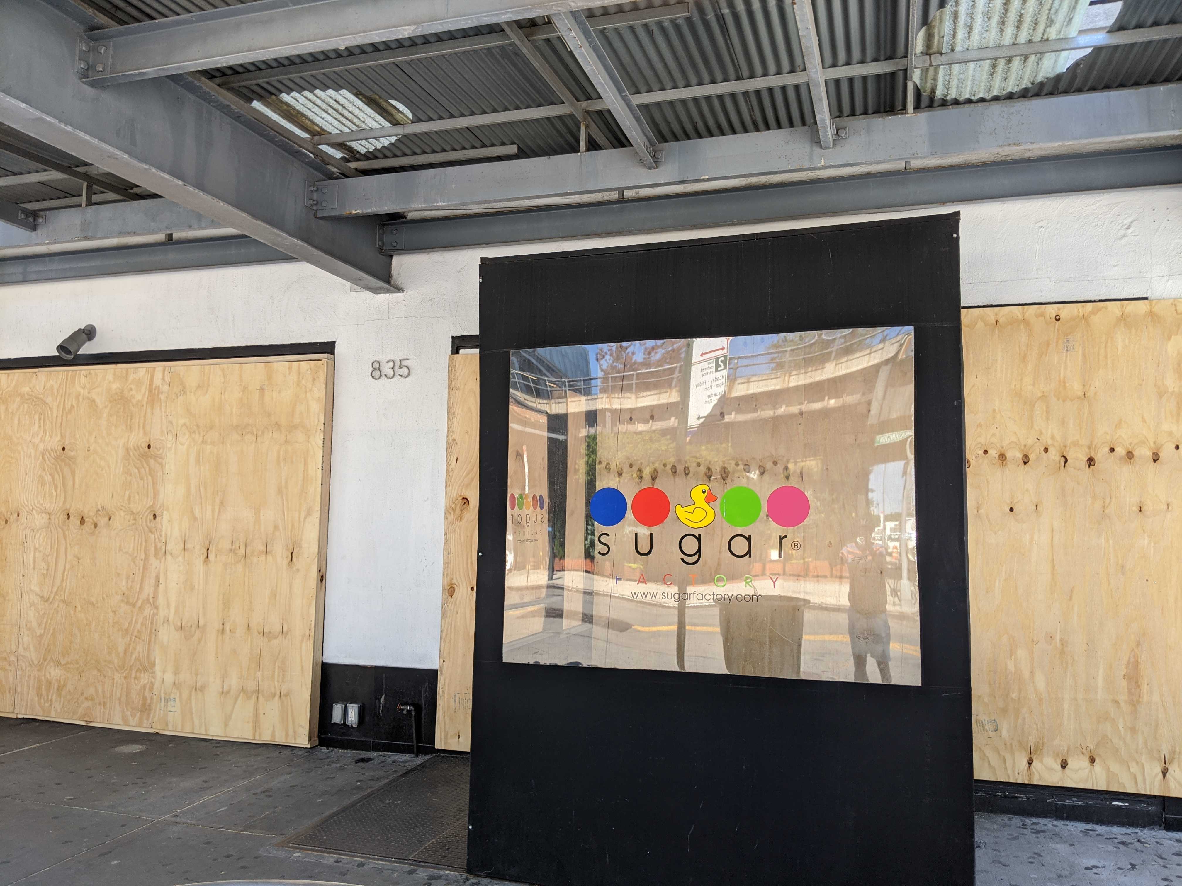 A storefront with wooden boards covering its windows. In front, a sign says “Sugar Factory” with a colorful logo