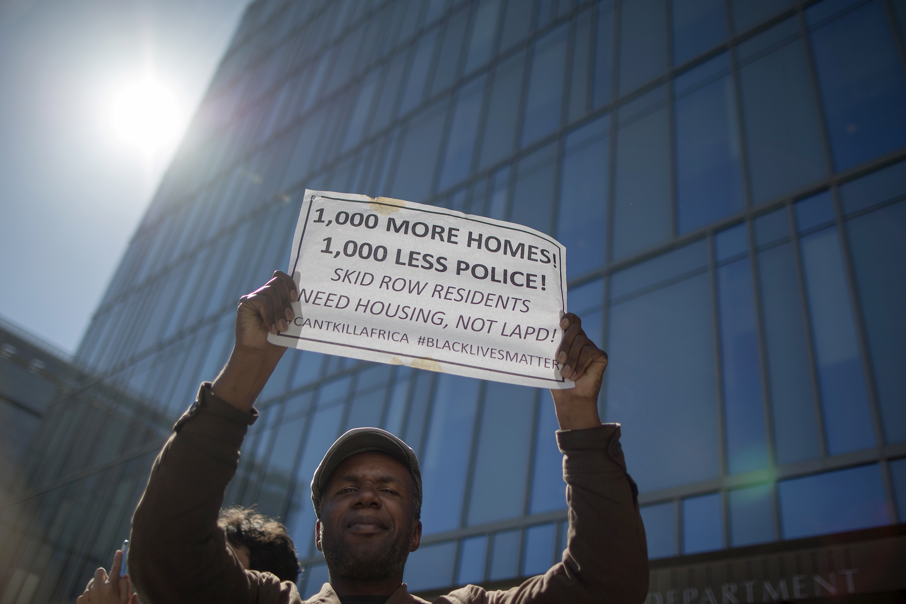 A protester standing in front of a glass skyscraper is holding a sign that says “1,000 more homes, 1,000 less police.”