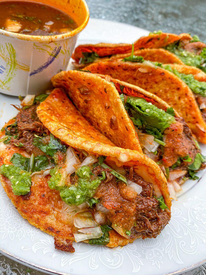 A close-up shot of a birria taco, which is stewed meat served in a taco. A bowl of the stew is sitting in the back of the photo
