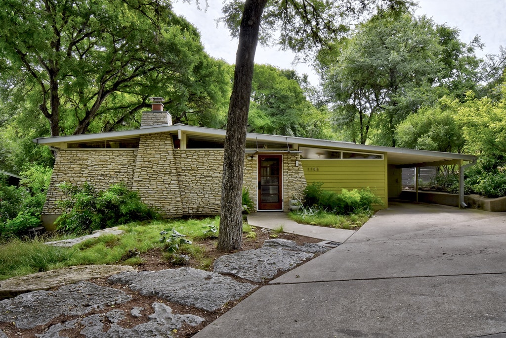 An exterior view of a midcentury modern house in Austin. There is a concrete driveway, a stone wall, and a green exterior wall. 