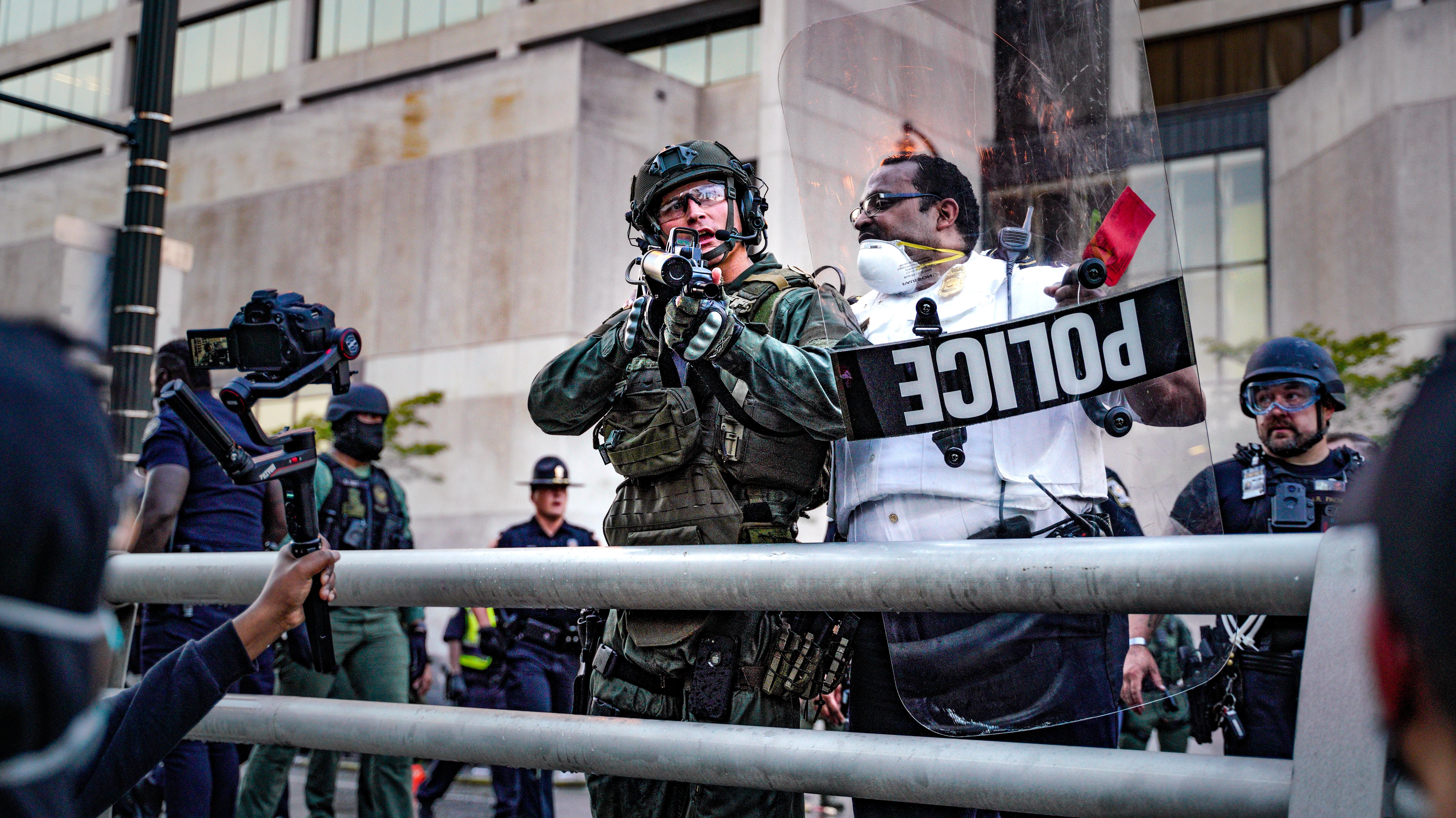 A protester holds a camera in front of a police officer in riot gear.