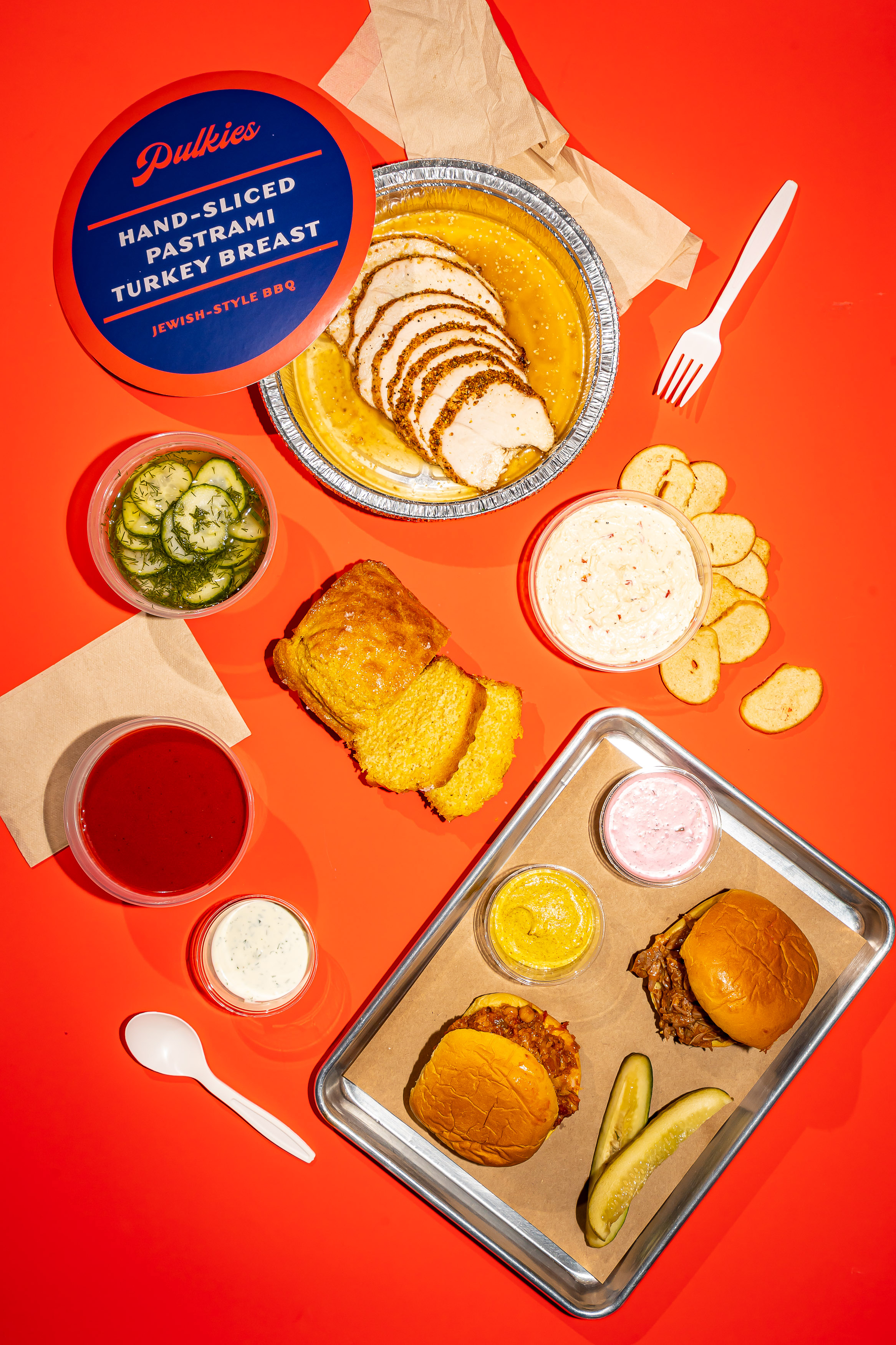An overhead photograph of a spread of foods, including two pulled turkey sandwiches, plastic to-go containers of dips, sliced breads, pickles, and plastic utensils