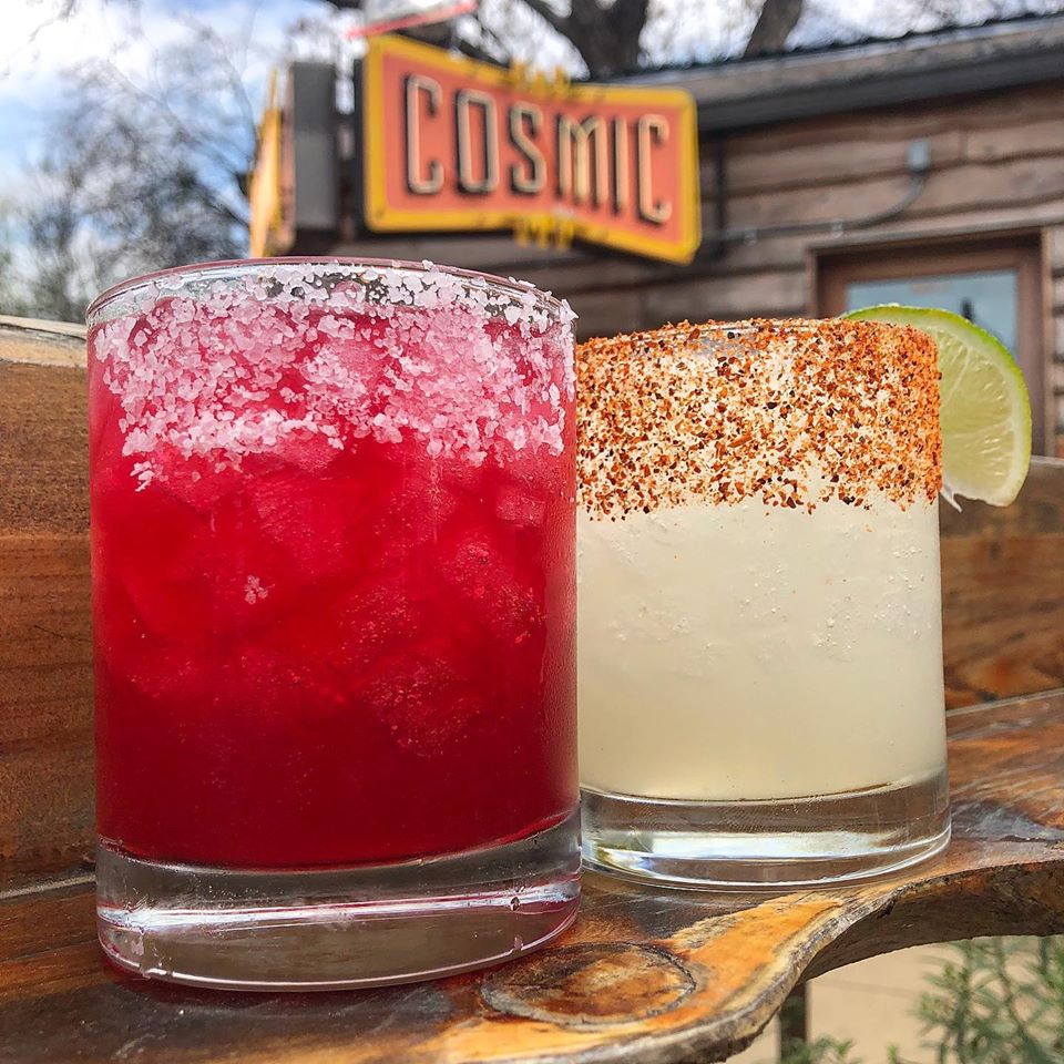 A red margarita with white salt and a green margarita with spicy salt on a wood table with the cosmic sign in the background
