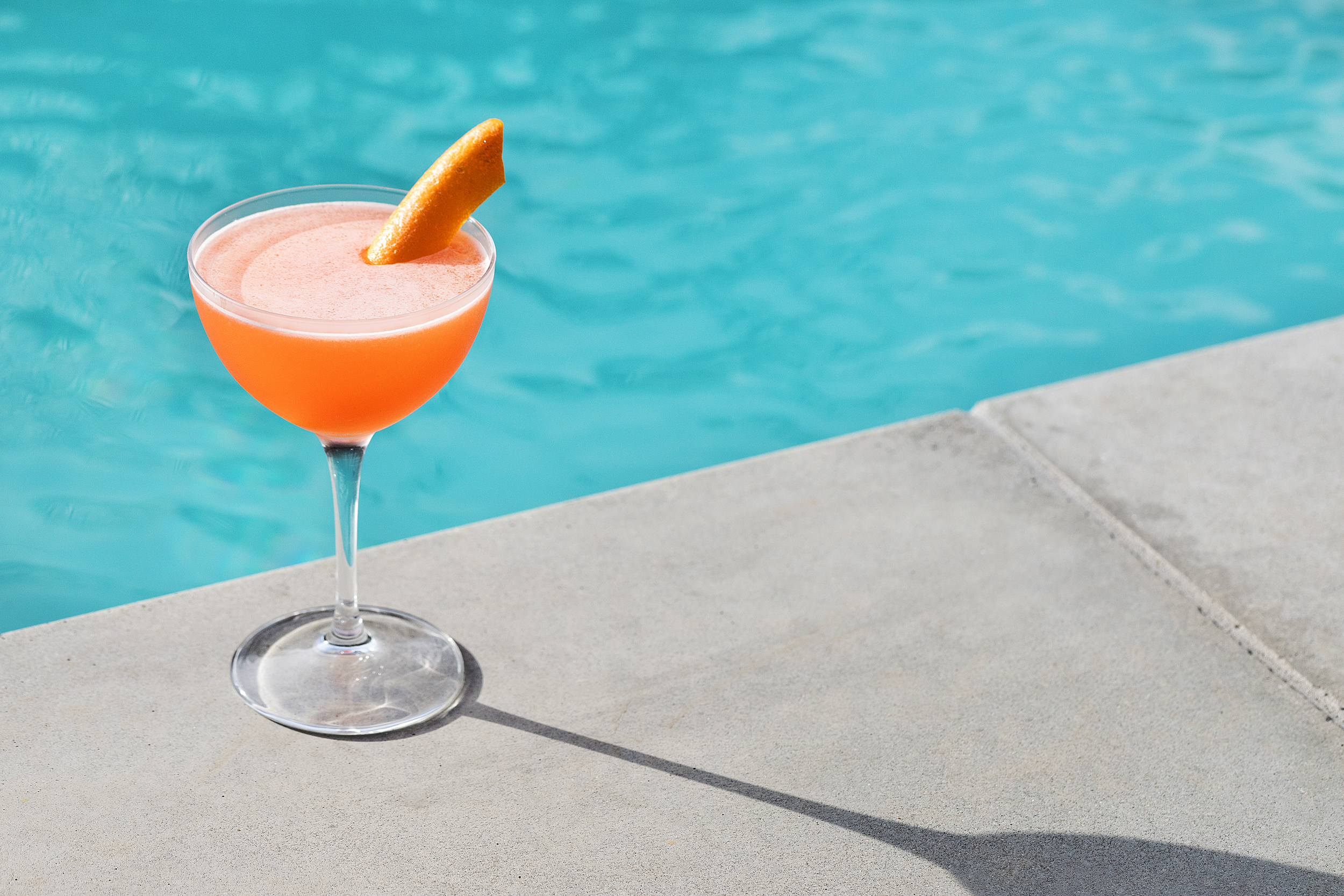 An orange cocktail in a tall glass sits next to a shimmering pool.