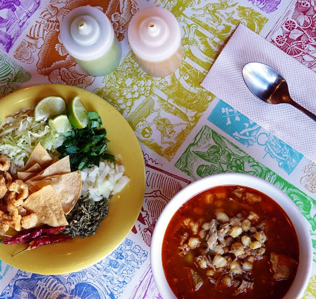 On a placemat, a bowl of pozole sits next to a plate of accompaniments like limes and onions. 