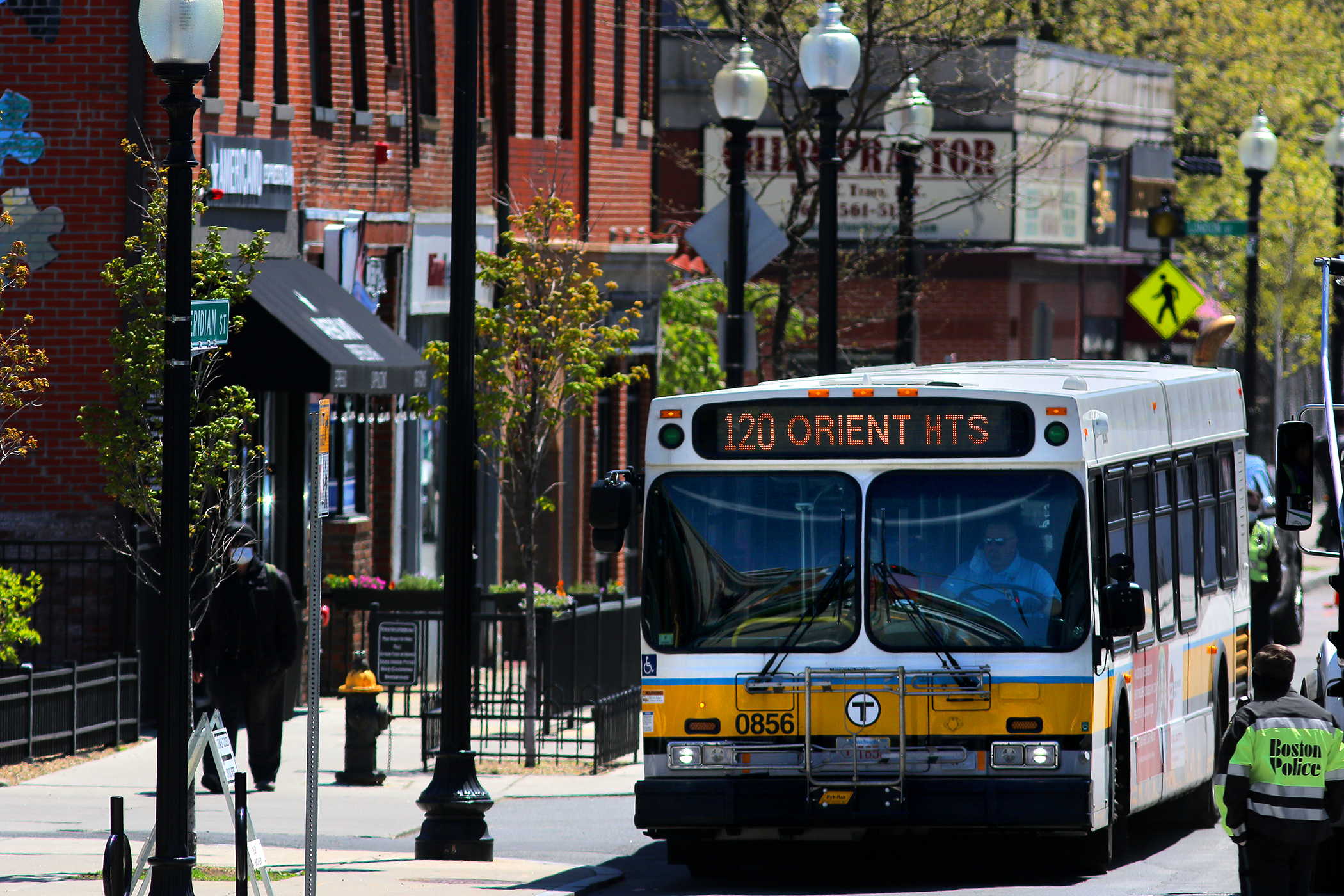 A yellow and blue striped bus with a heading that reads 120 Orient Heights travels along Meridian Street in East Boston, a busy street lined with brick buildings.