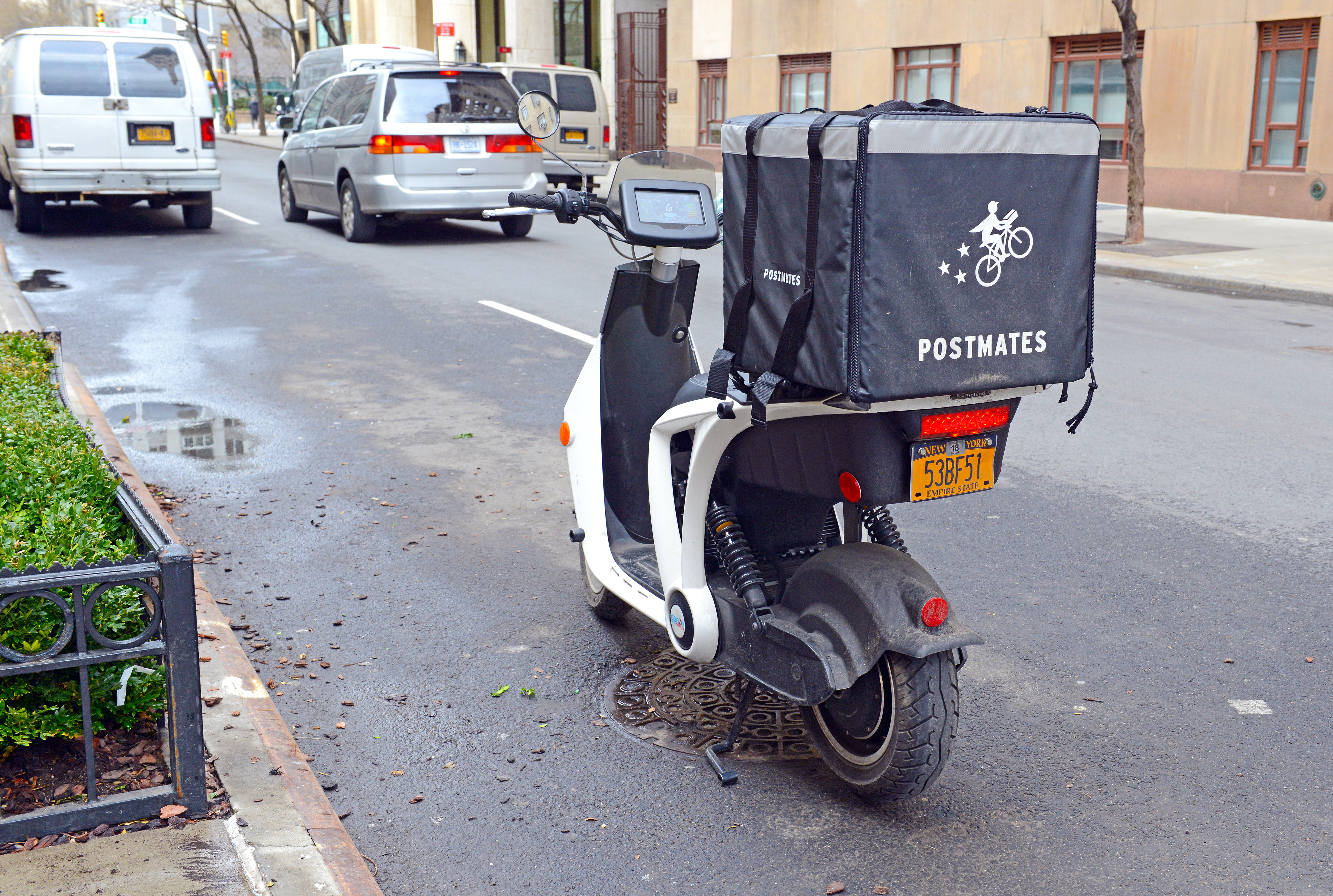 A motorbike parked on the street with a Postmates delivery bag attached to the back.