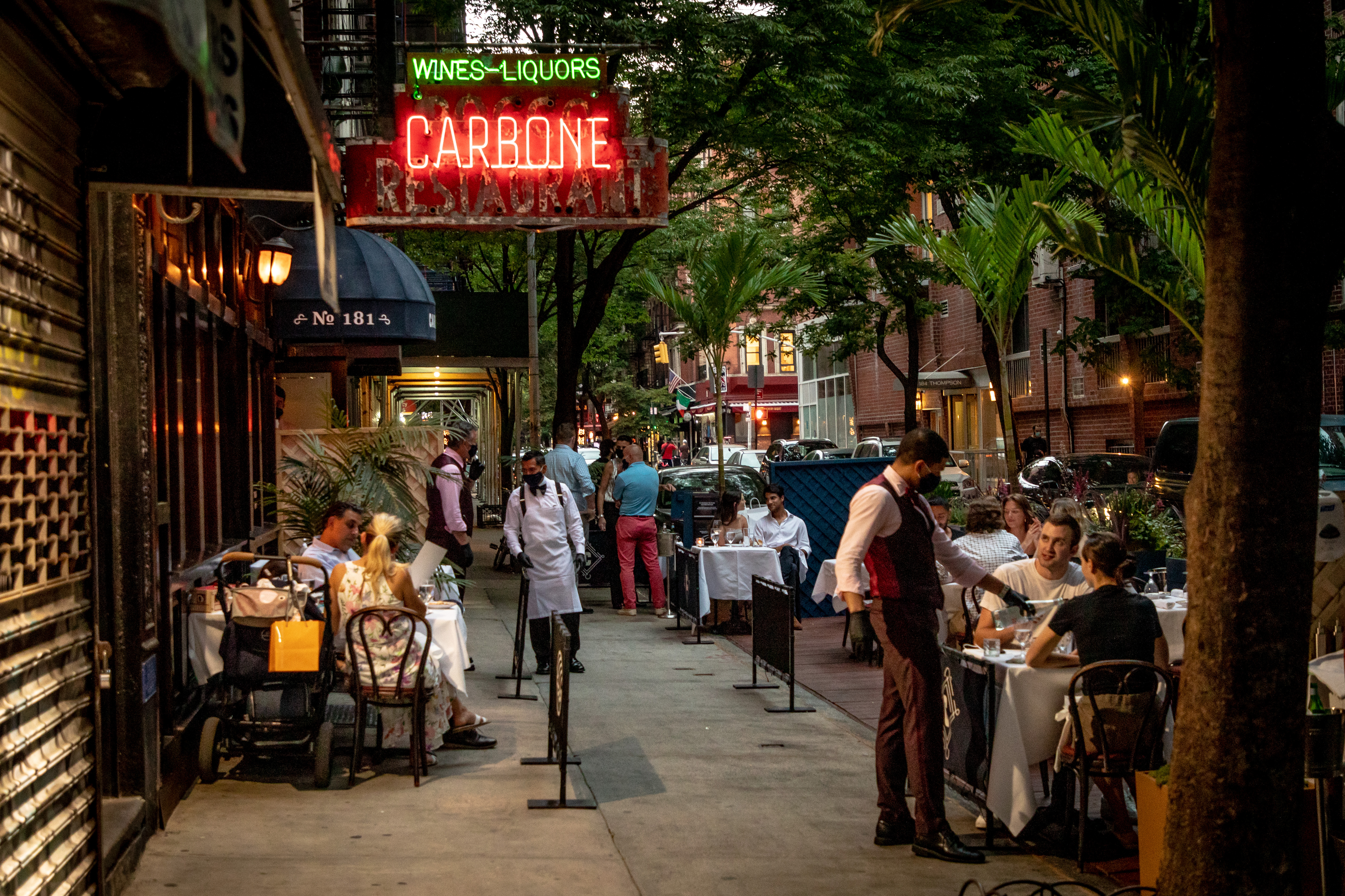 Waiters attend to groups of diners sitting at outdoor tables on a tree-lined street. To the left, a neon illuminated sign “Wines Liquors Carbone” shines.
