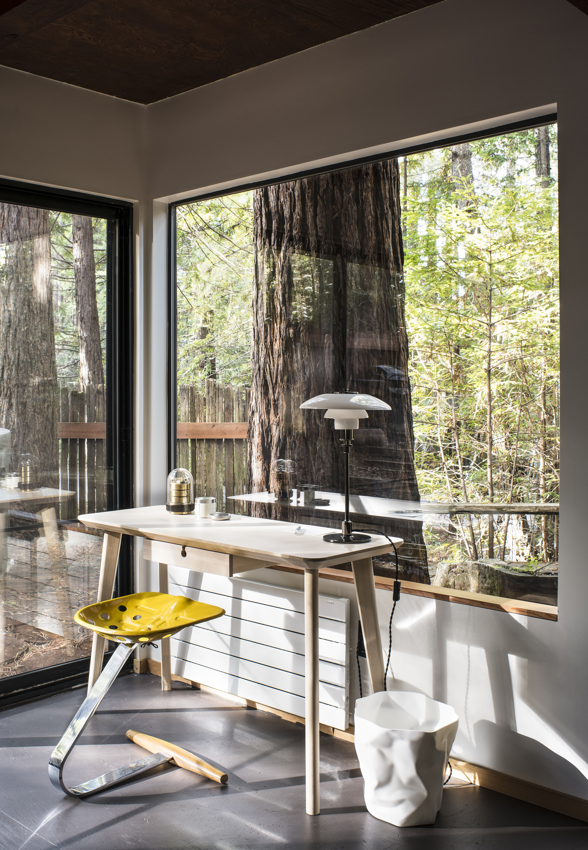 A white desk with a yellow metal stool in front of a large window with views to a redwood forest