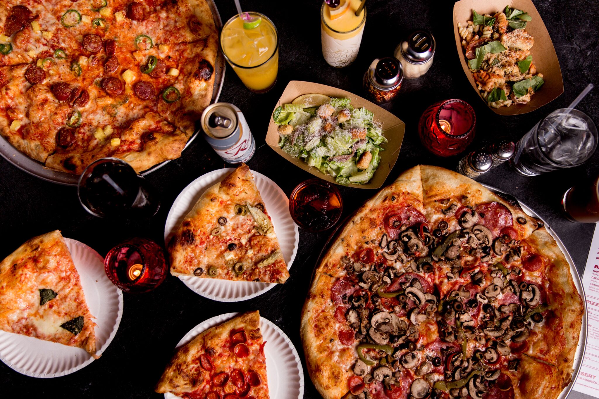 A top-down view of several different pizzas, with a variety of toppings, alongside cocktails, beers, and salads.