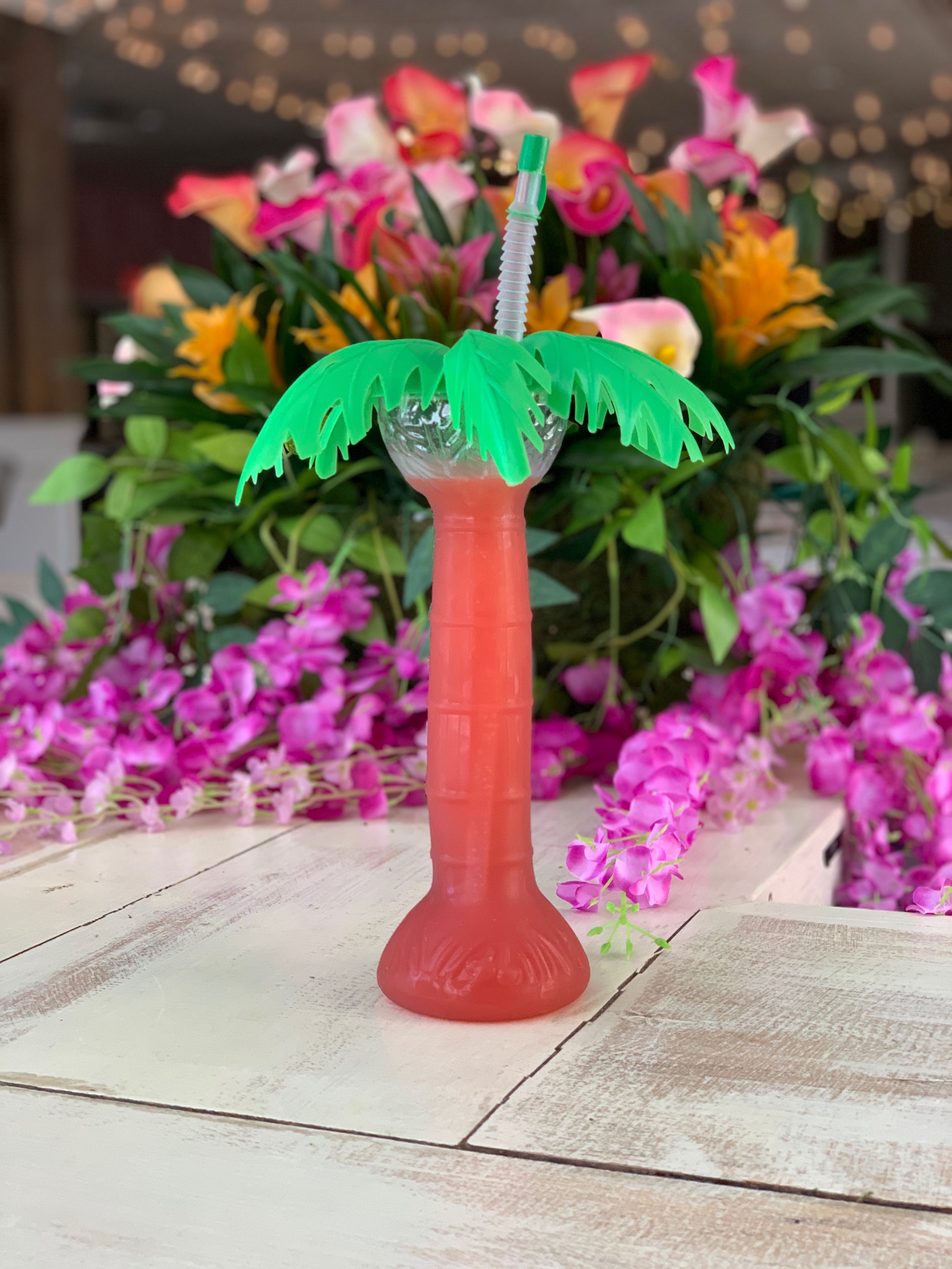 Plastic palm tree cup full of a pink frozen cocktail sits on a table in front of a colorful floral arrangement