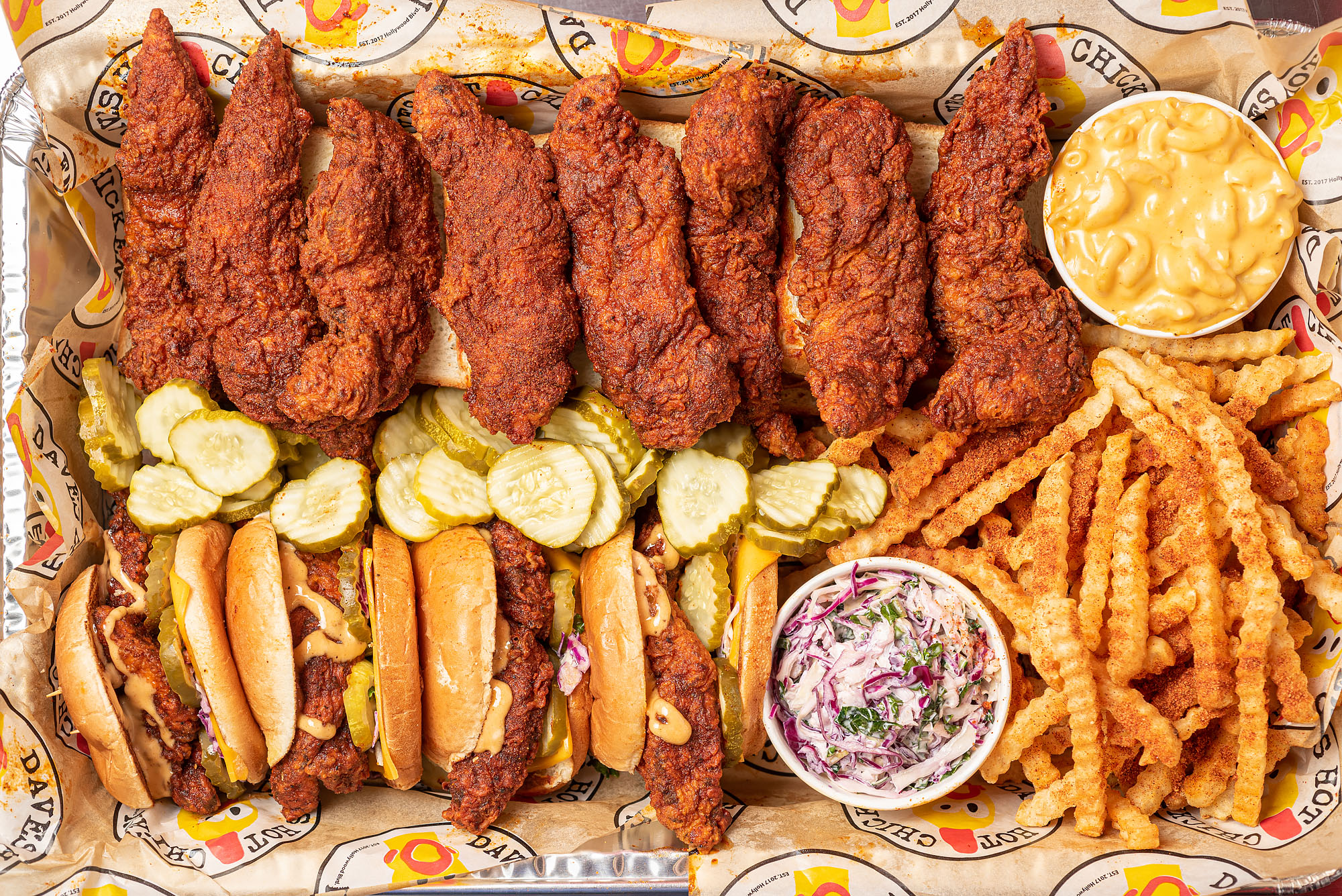 Hot chicken tenders on a tray divided by pickle chips with hot chicken sandwiches on the other side, french fries, cole slaw, and macaroni.