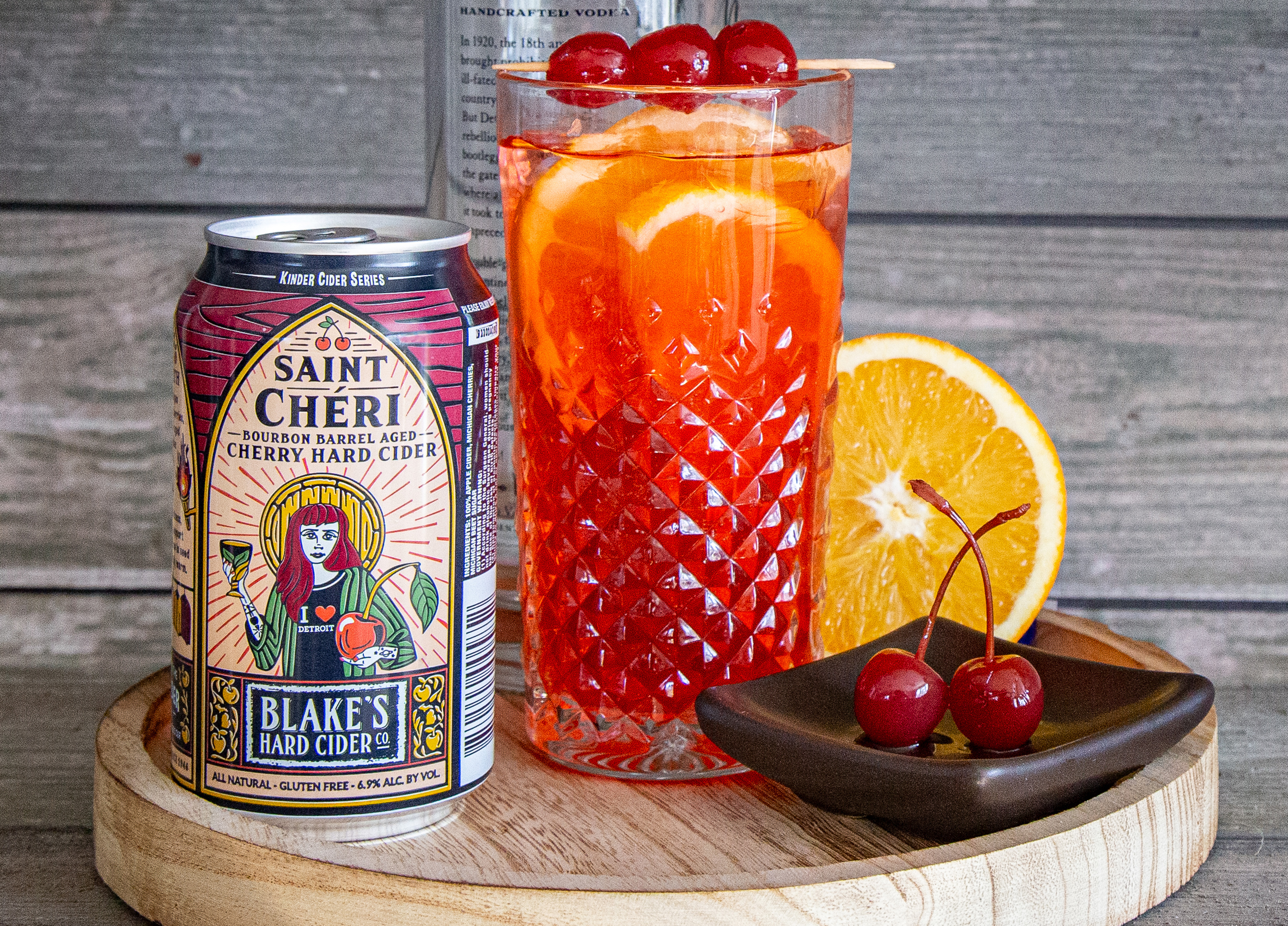 Saint Cheri cider in a red can next to a glass of cherry cider and a plate with two maraschino cherries. 