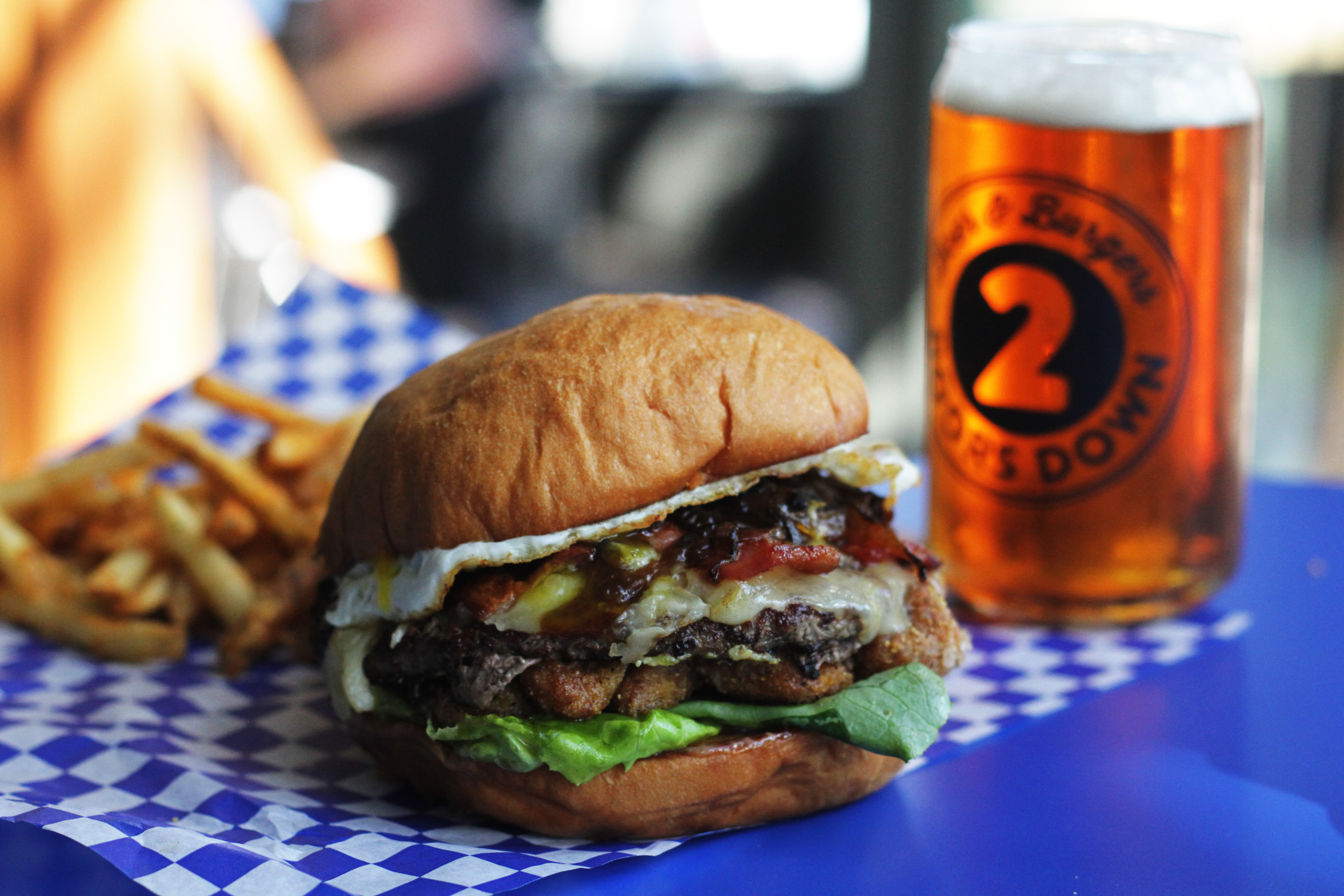 A burger loaded with toppings at Two Doors Down, next to a full glass of beer and fries in the background