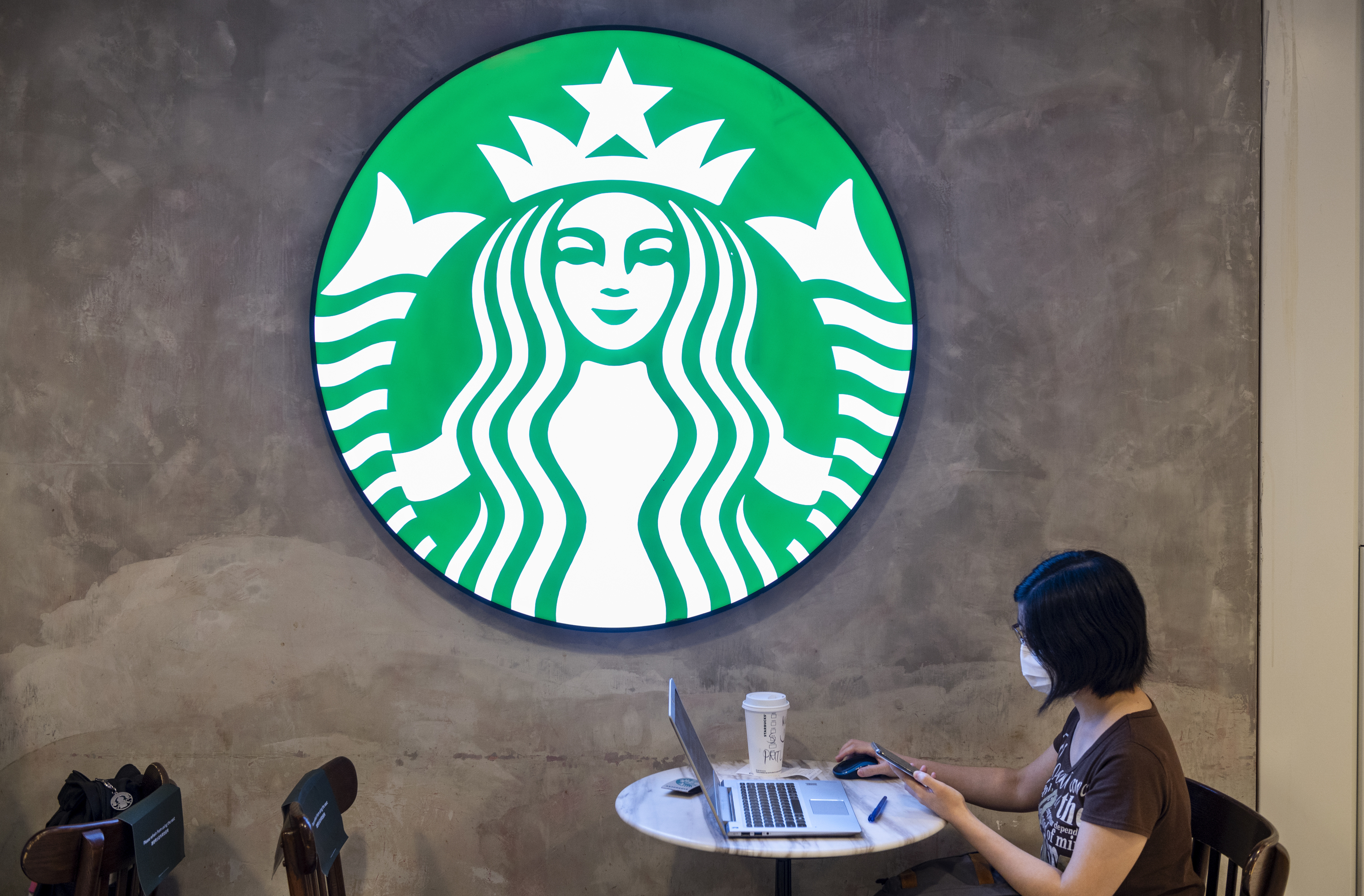 A masked woman with short dark hair sits at a marble cafe table, working on her laptop under an illuminated Starbucks logo.