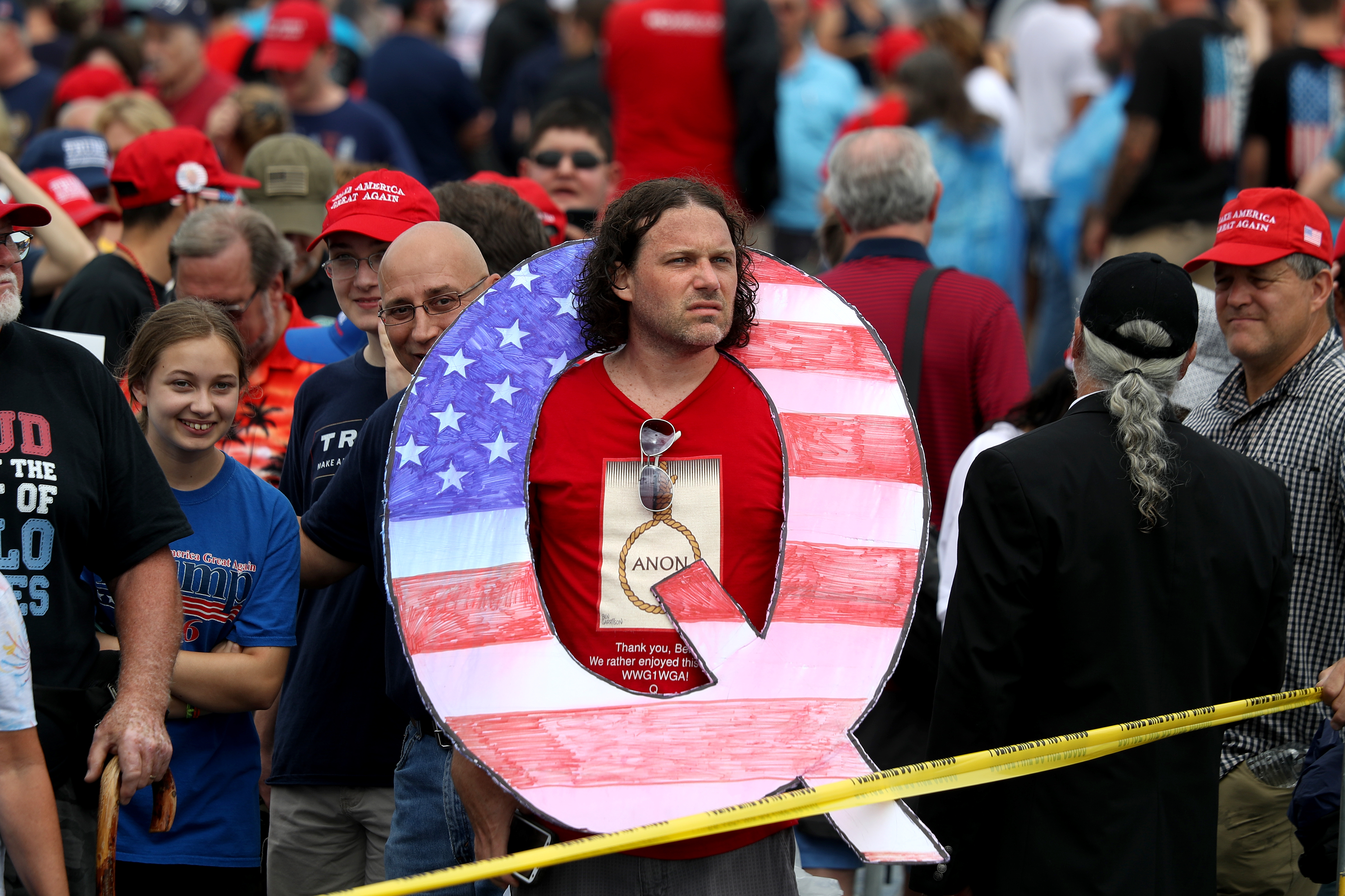 Trump rally attendee wearing a red shirt holds a large red, white, and blue “Q” sign while waiting in line on to see President Donald J. Trump at his rally August 2, 2018. “Q” represents QAnon, a debunked conspiracy theory group.
