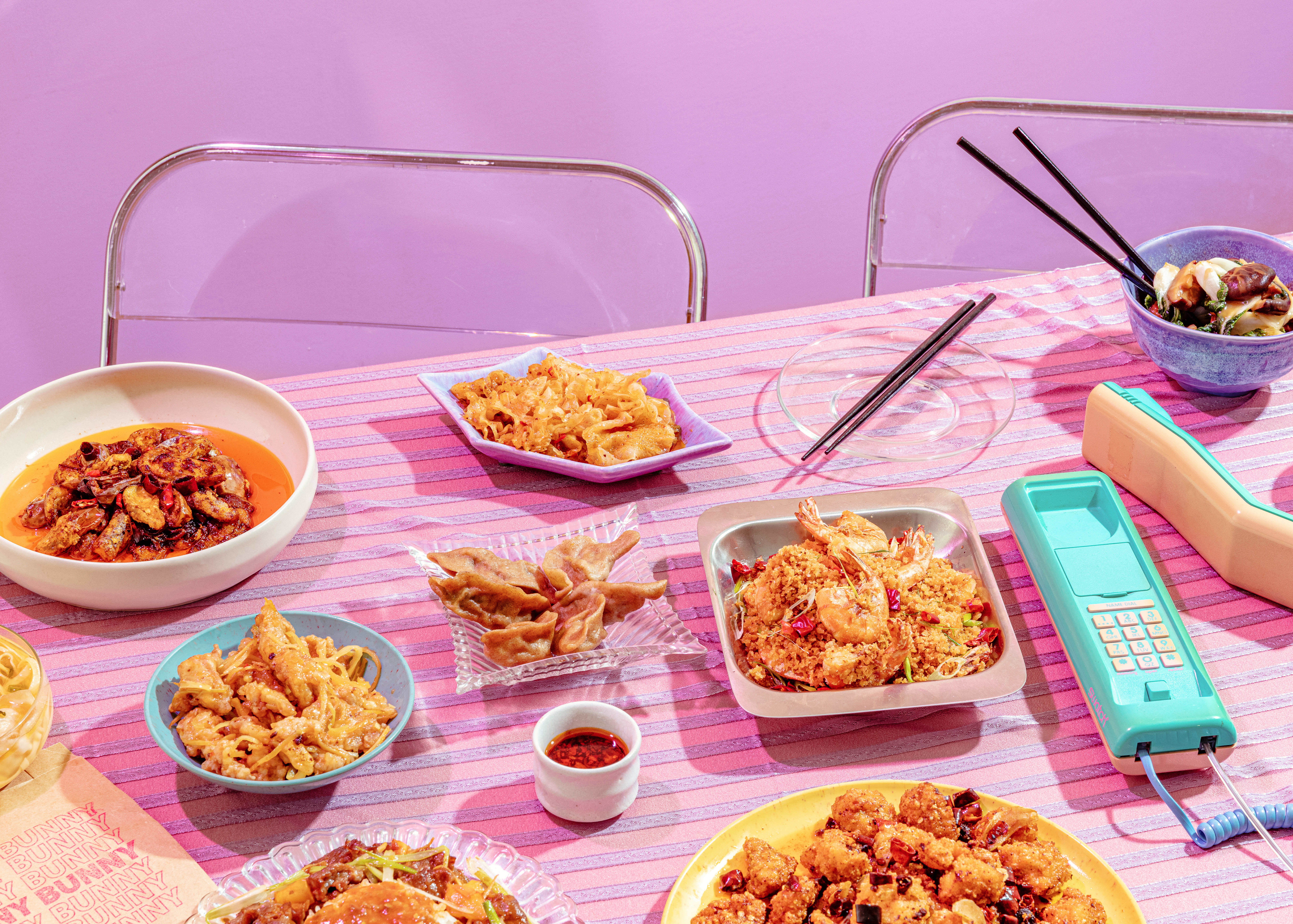 Dishes on a retro, 1980s-styled table with dishes from Bunny Bunny surrounded by silver and clear plastic chairs and a pastel-colored corded phone.