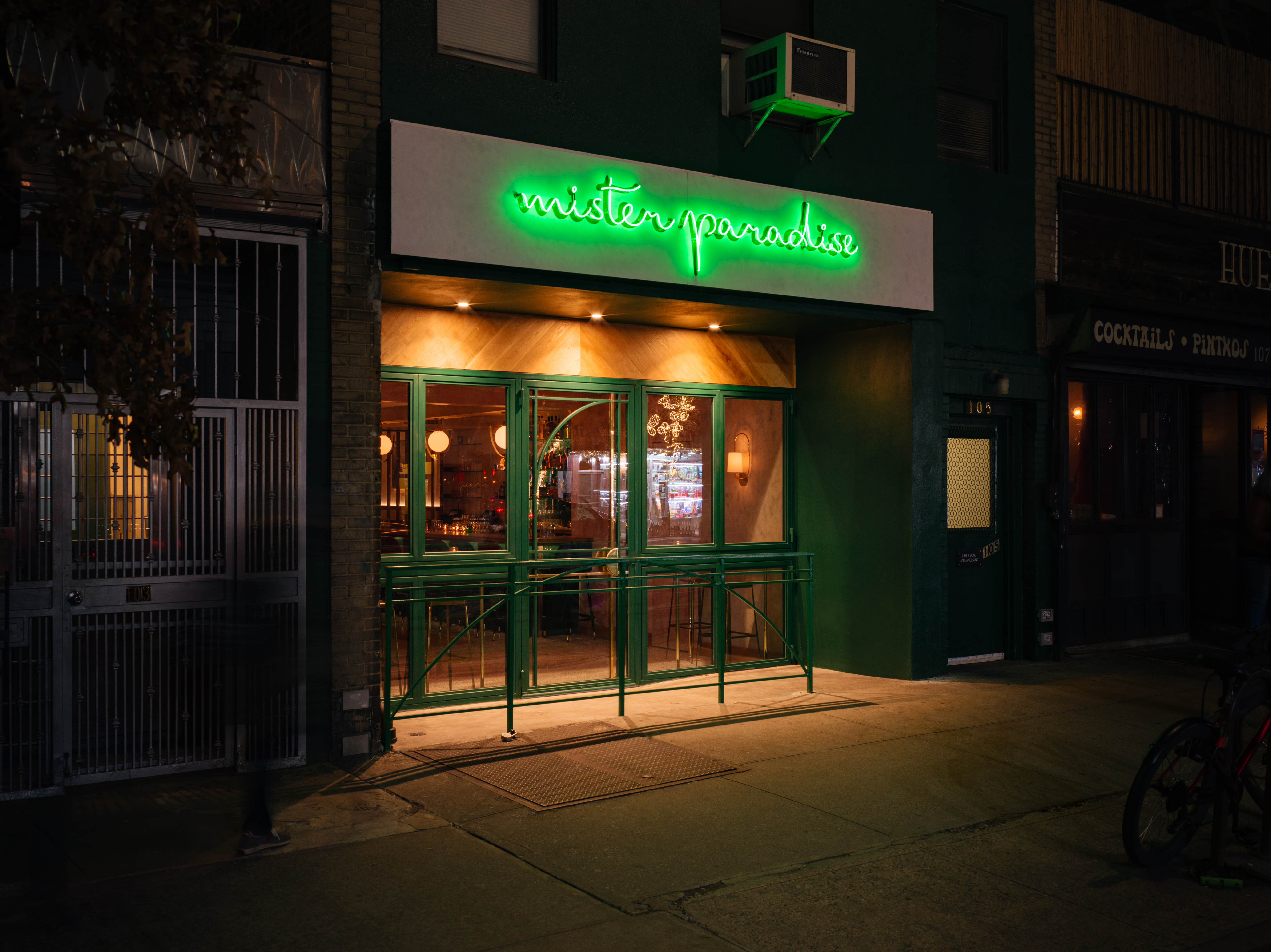 The exterior of a bar with a green railing and a green neon sign that says “Mister Paradise” in cursive lettering