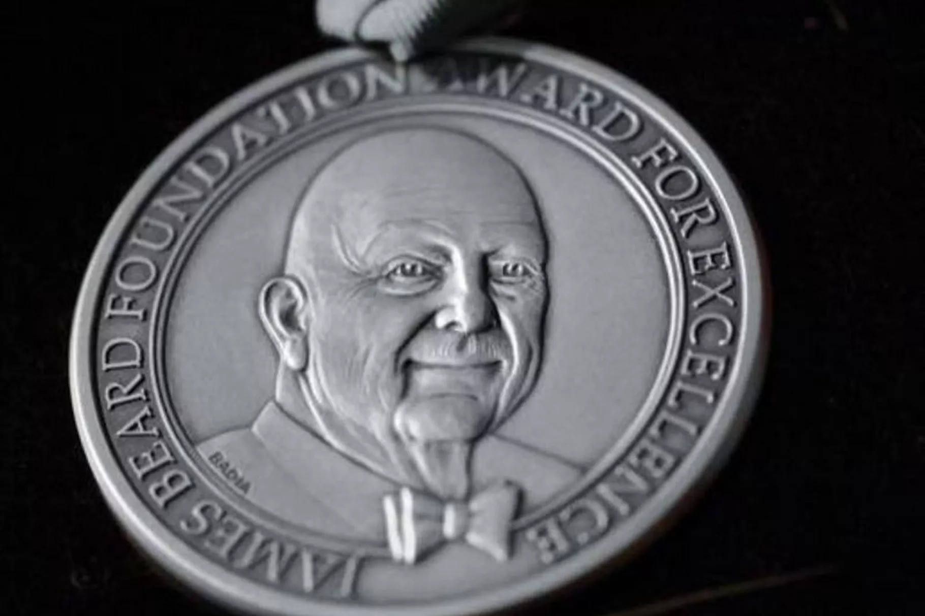 A silver James Beard Awards medal featuring James Beard’s face at center and the words “James Beard Foundation Award for Excellence” along the border.