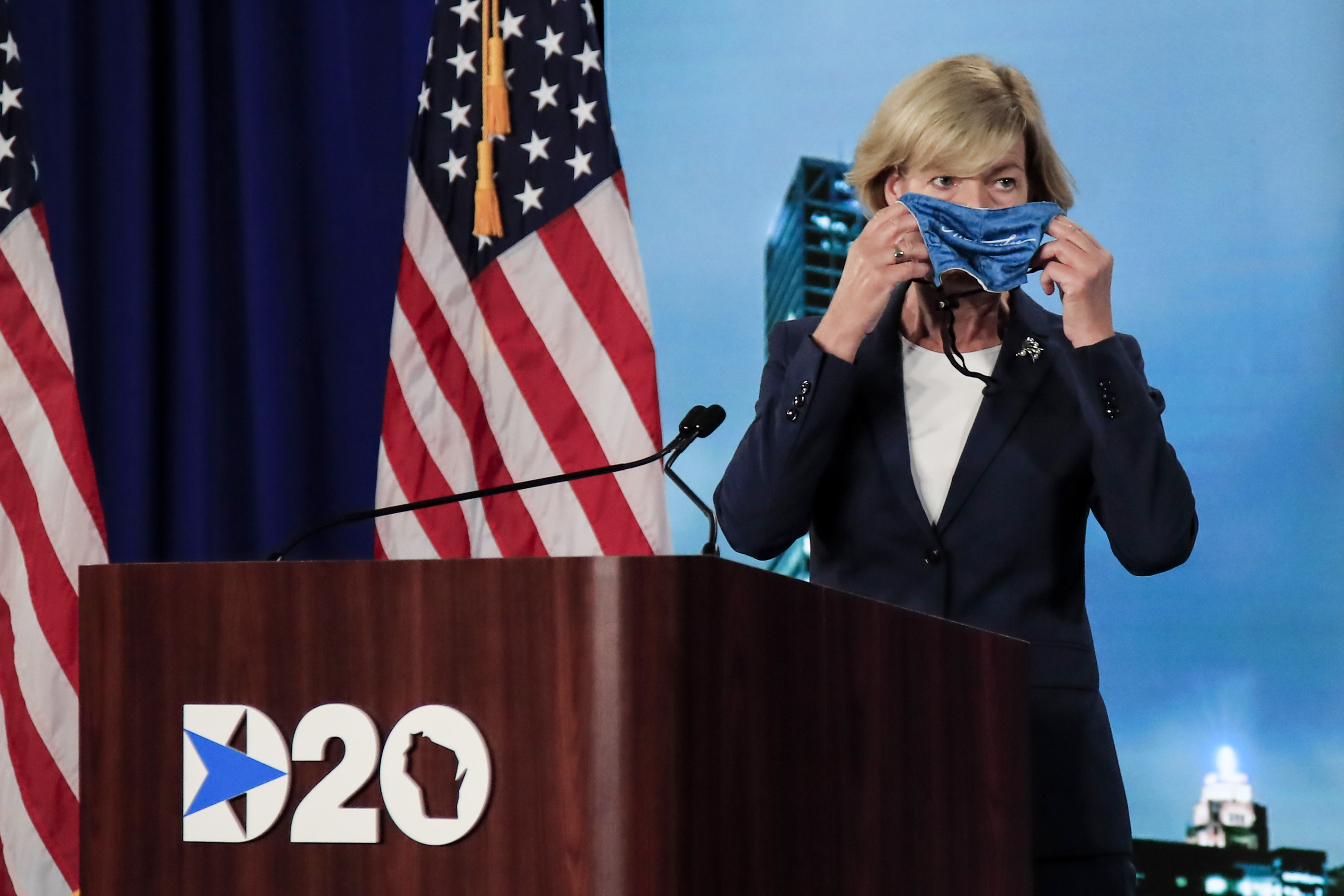 Senator Tammy Baldwin holding a mask to her face while at the Democratic National Convention podium.