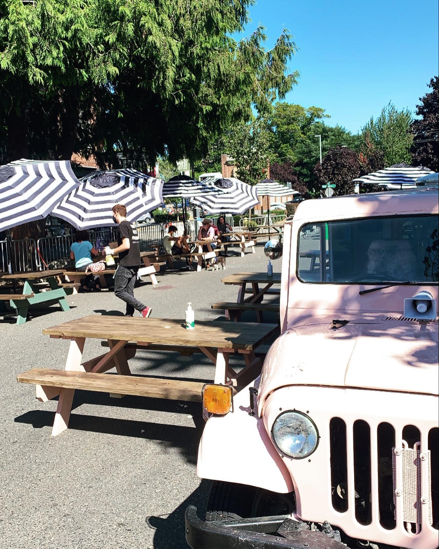 Tables are lined up on W Crockett Street in Queen Anne, with striped umbrellas and a masked server bringing food to customers. In the foreground is a pink jeep.