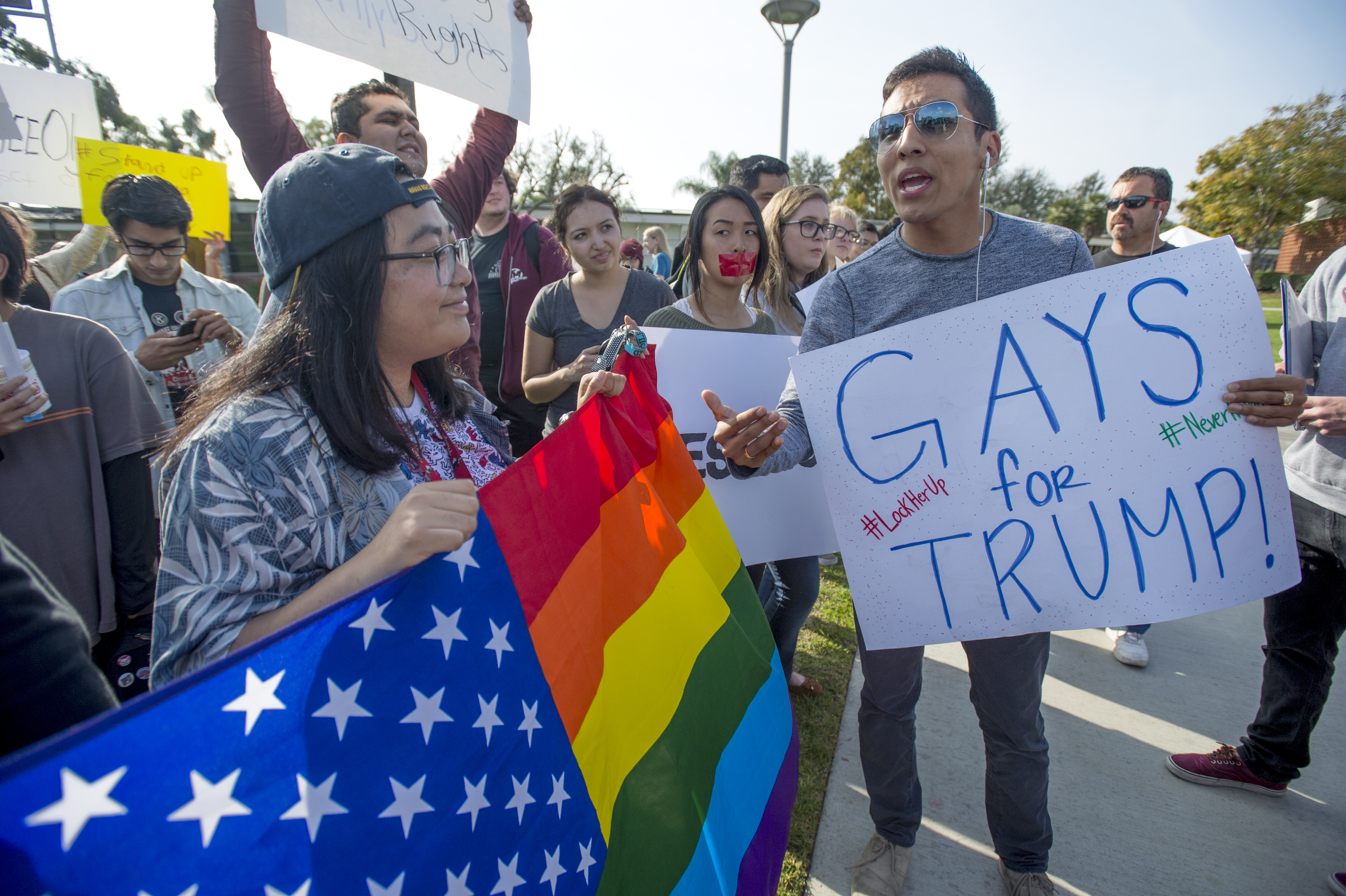 A protester carrying a rainbow-striped American flag looks at a counterprotester carrying a sign that reads, “Gays for Trump.”