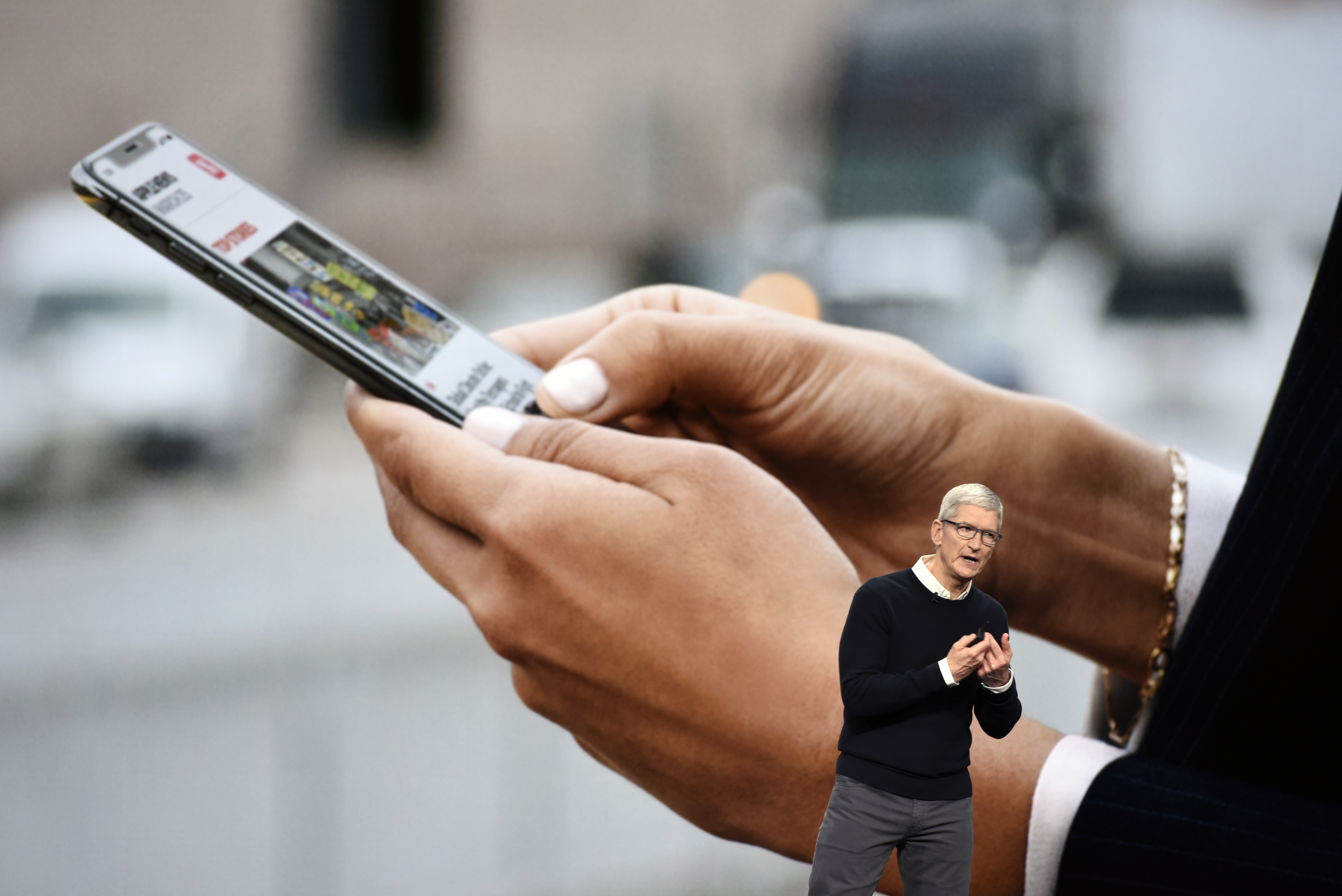Apple CEO Tim Cook onstage in front of a wall-sized screen showing two hands holding an iPhone.
