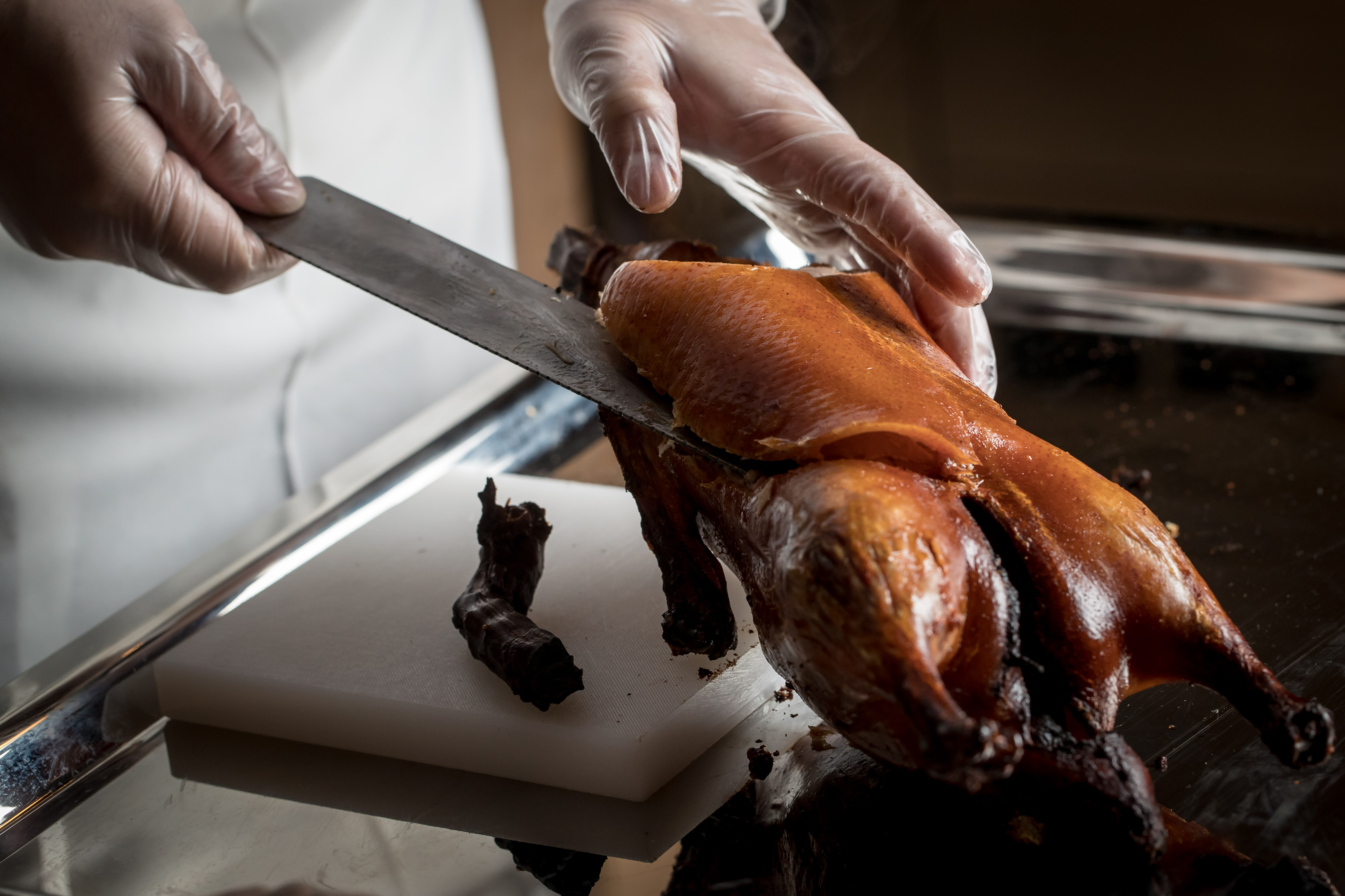 A chef’s gloved hands reaching towards a roast duck on a table. One hand holds a knife which is propping up the duck