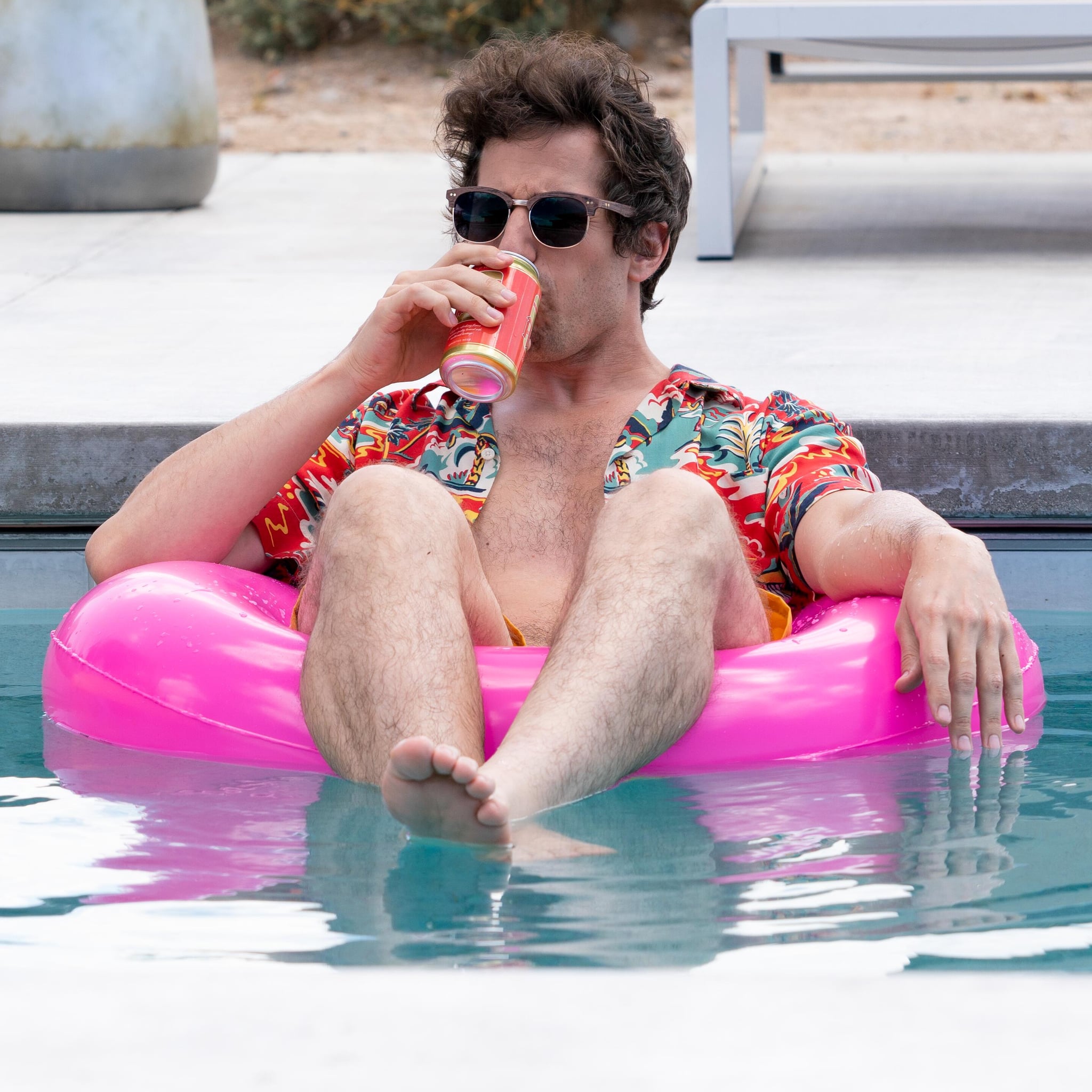 A man wearing sunglasses and a Hawaiian shirt floats in a pool drinking beer.