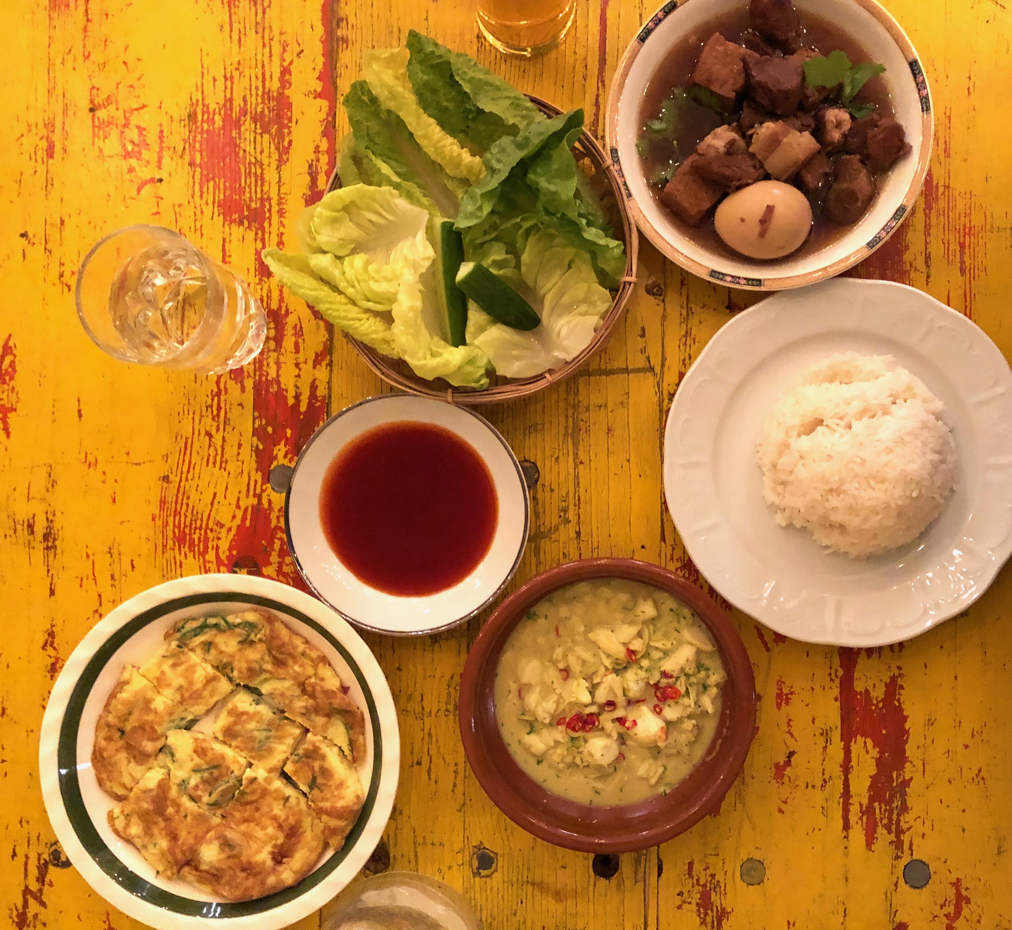 A spread of dishes, including pork belly in a dark soy sauce, green lettuce leaves, a mound of white rice, and a sliced omelet sit over a burnished wood table