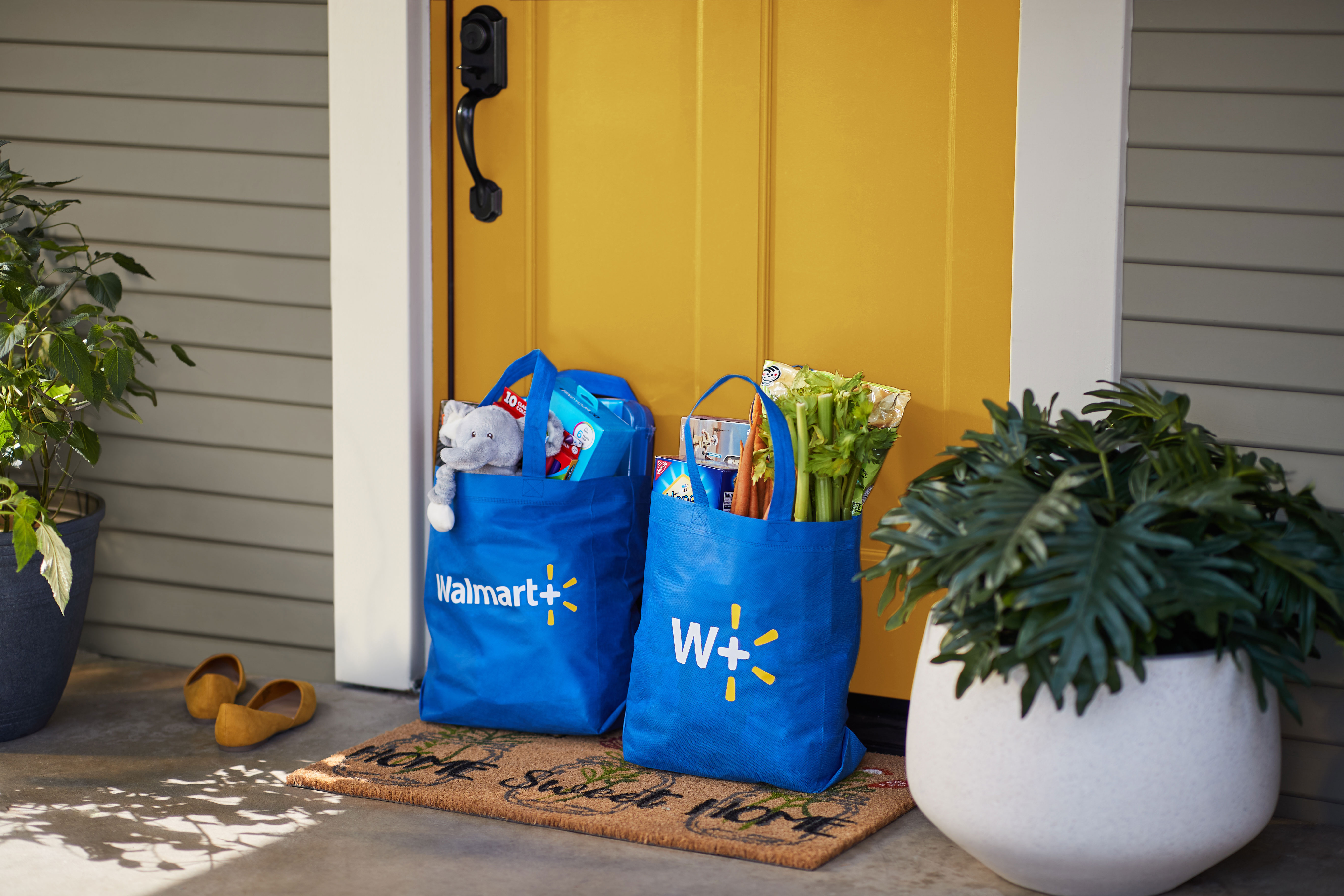 Two Walmart+ bags filled with groceries and other merchandise sit in front of the front door of a house.