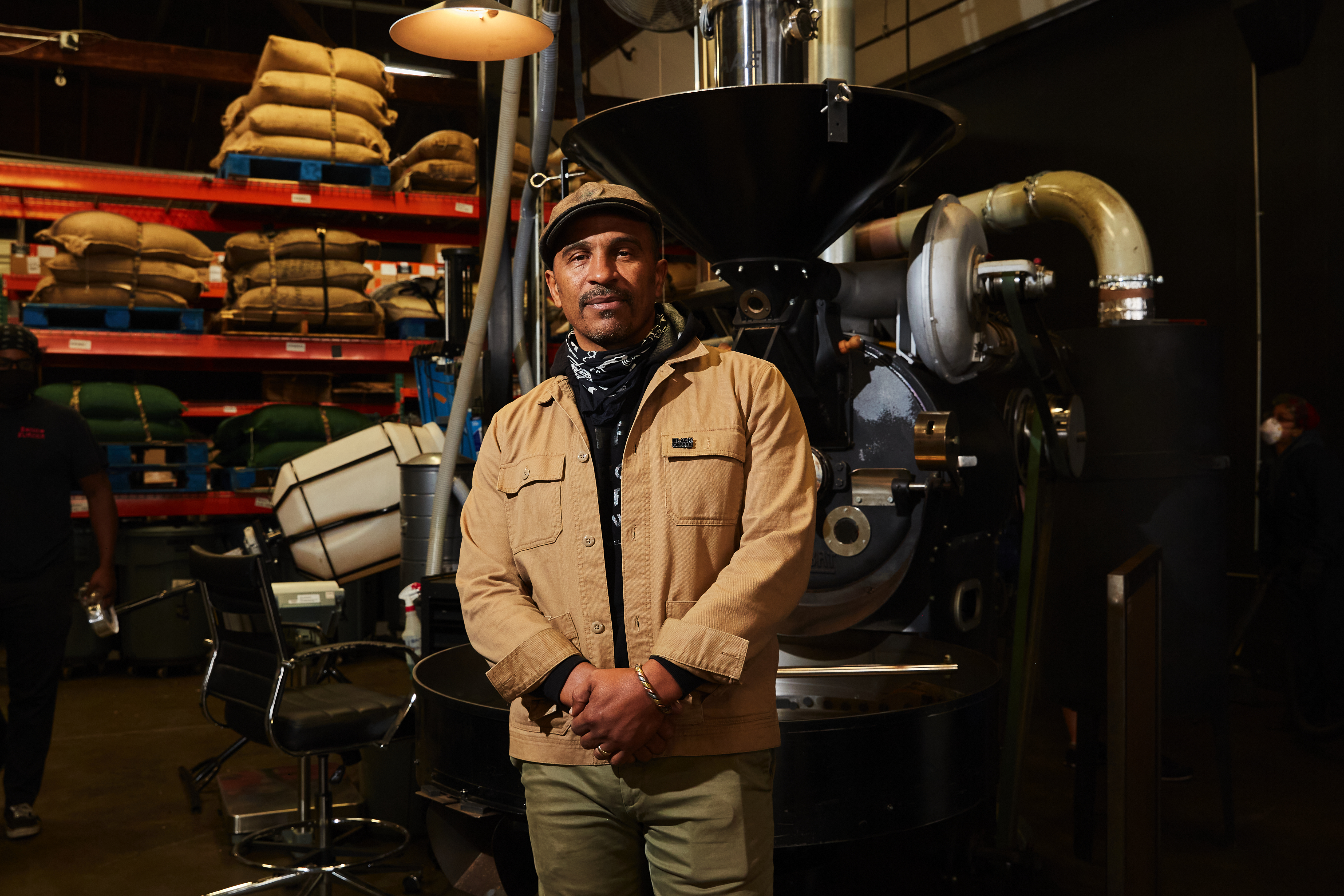 Man wearing cap standing in front of a coffee roaster, with bags of beans stacked on shelves in the background.