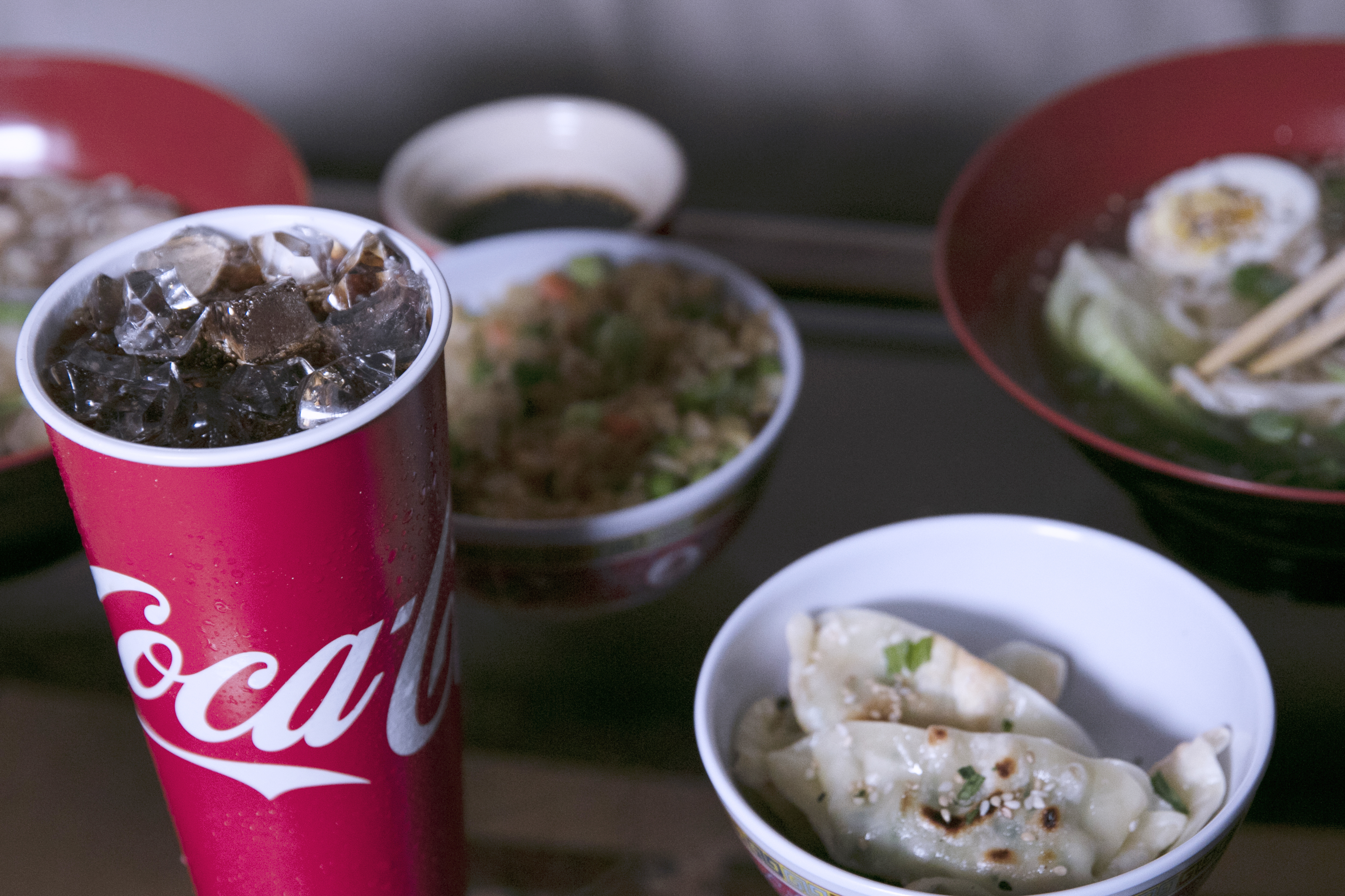 A paper cup of Coca-Cola on ice, sitting on a table with a variety of foods, such as dumplings and fried rice.