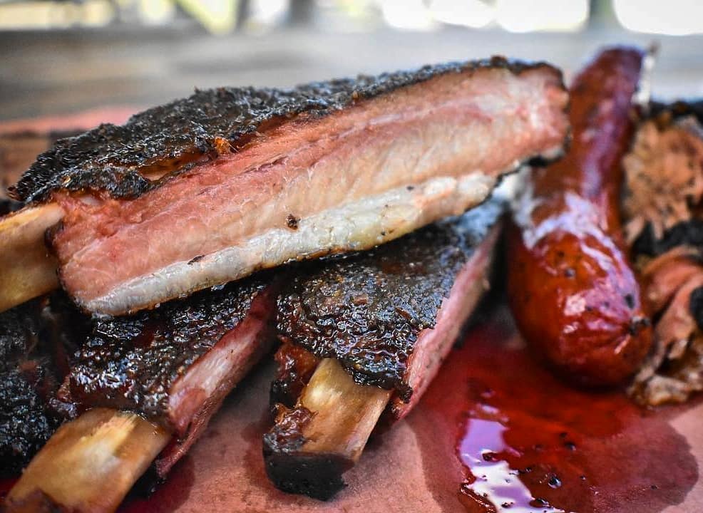 Close up of ribs stacked on greasy red butcher paper next to a link of sausage