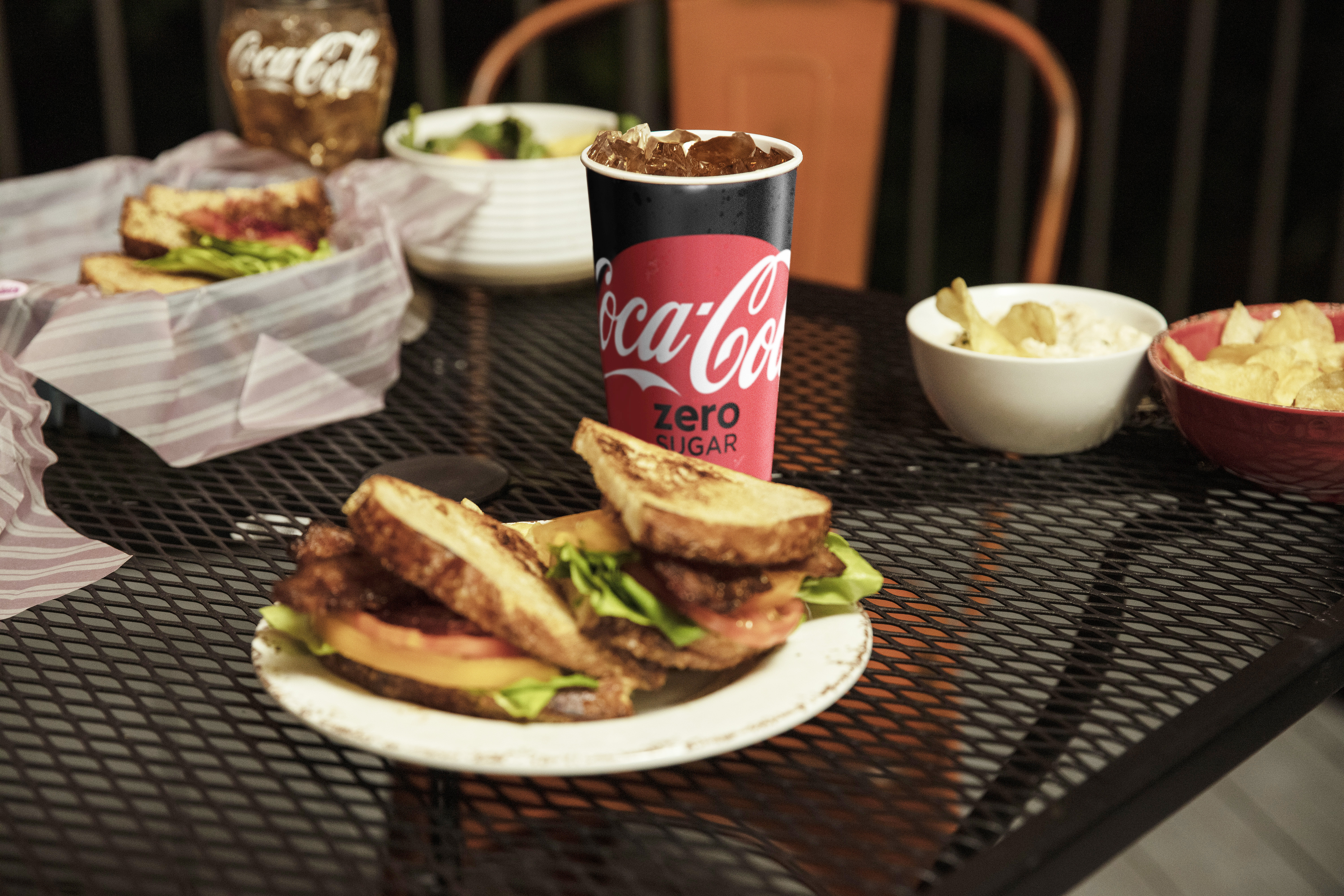 A dinner table topped with sandwiches, chips, and glasses of Coca-Cola soda.