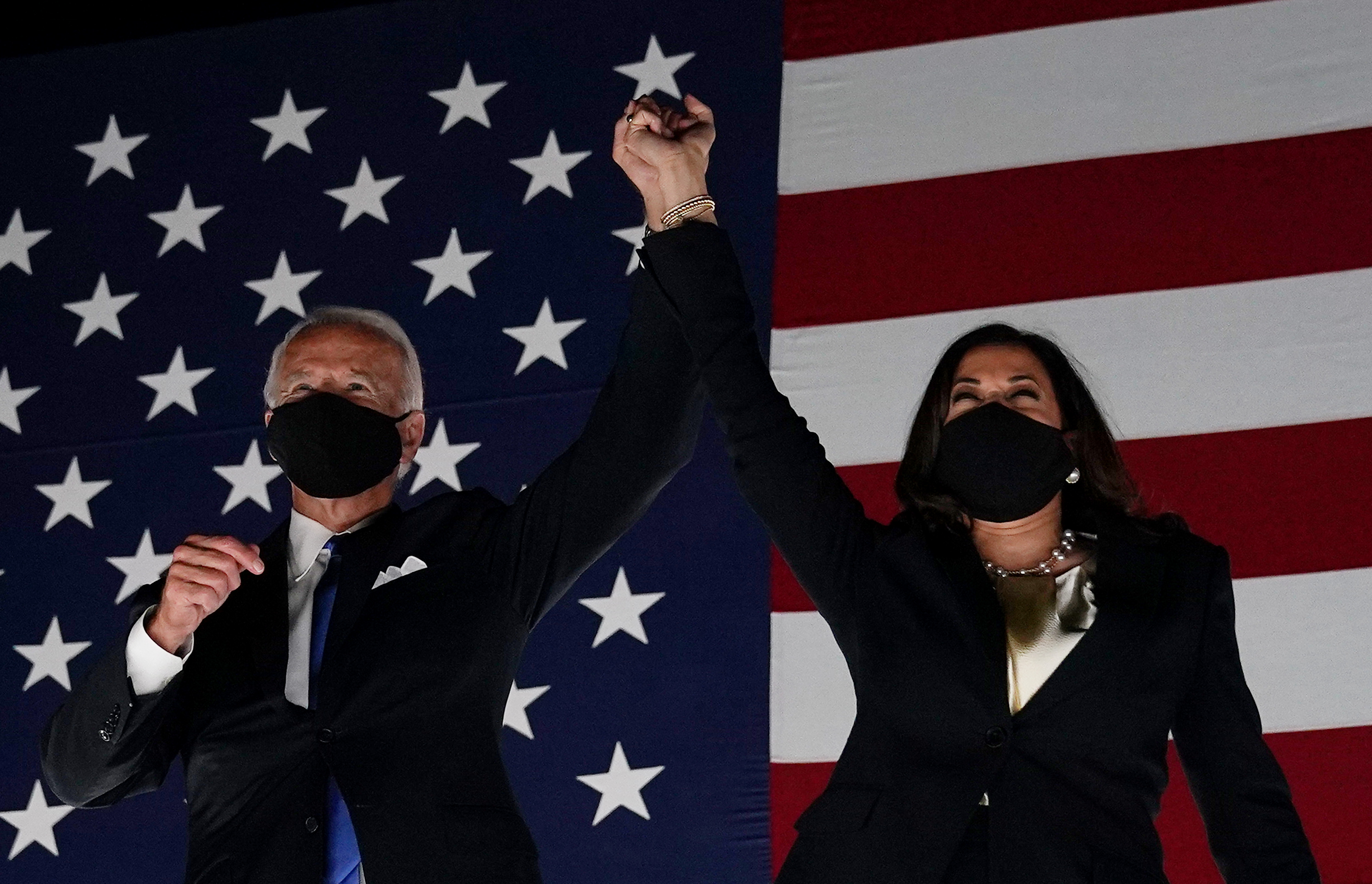 Joe Biden and Kamala Harris raise their held hands in front of a wall-sized American flag backdrop.