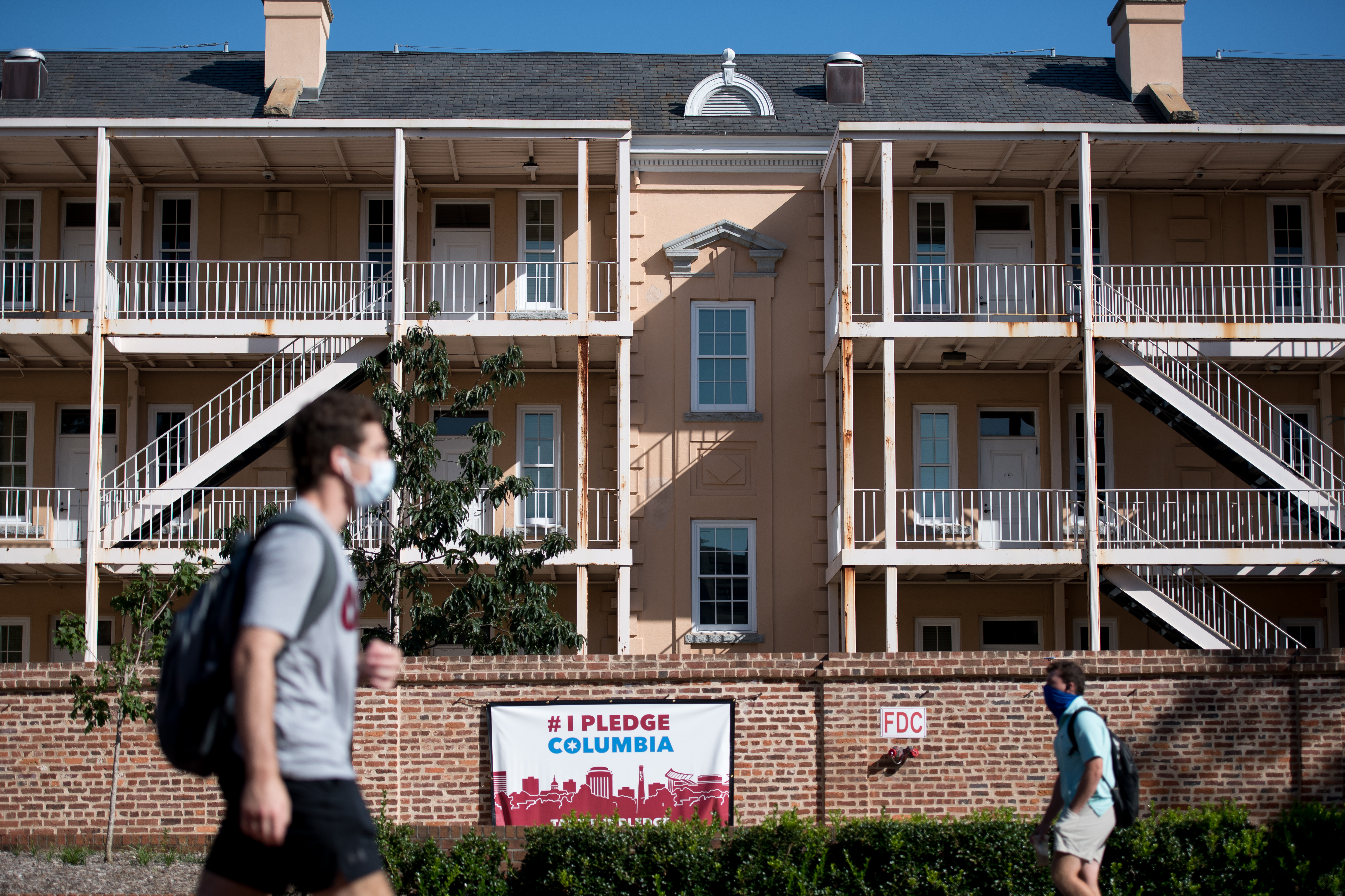 Two students wearing face masks walk past an apartment building on campus at the University of South Carolina, far apart from one another. There is no sign of activity in the apartment building.