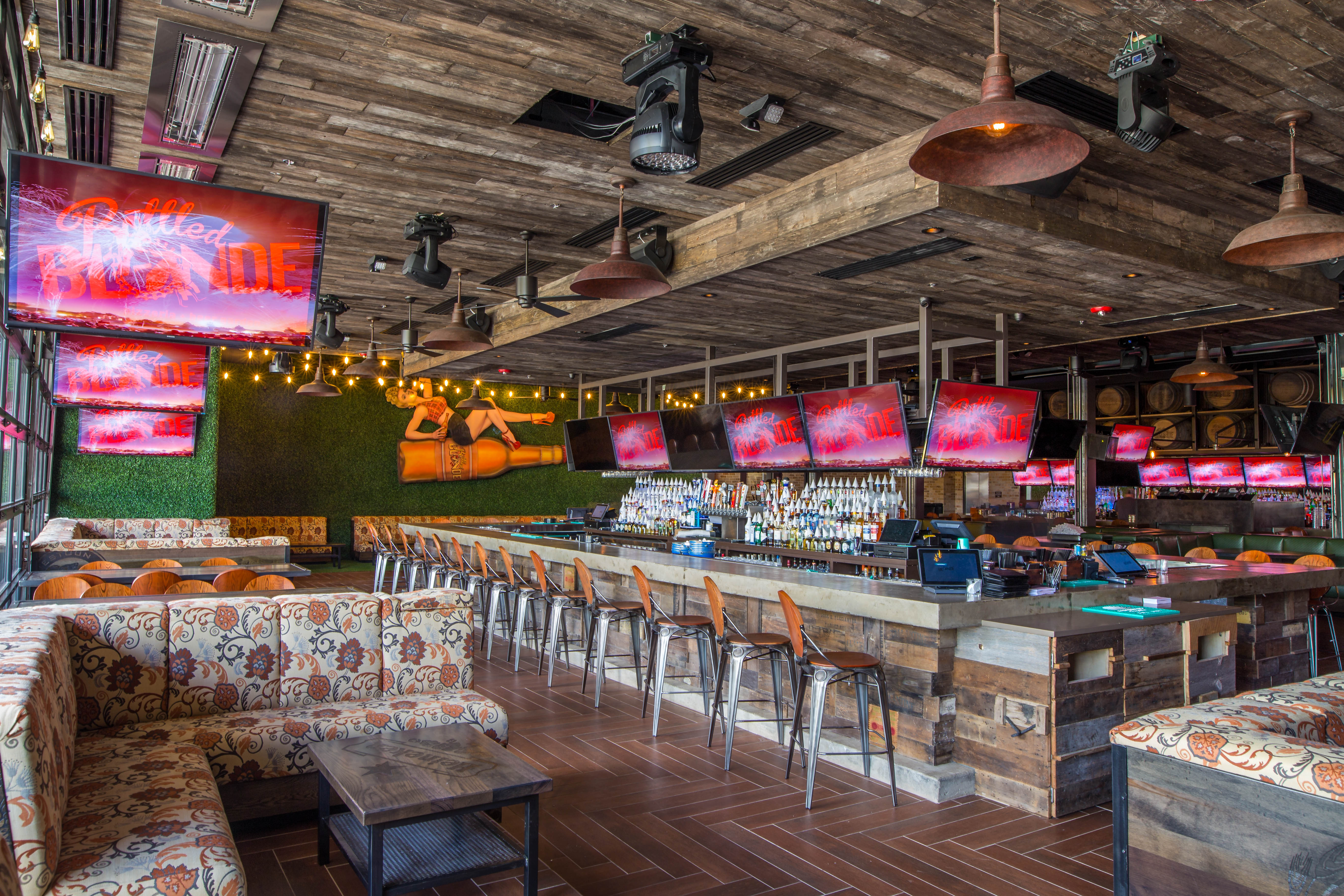 The interior of Bottled Blonde in Houston. Floral patterned banquettes sit on a herringbone pattern wood floor with a rustic wood bar and TV screens in the background.