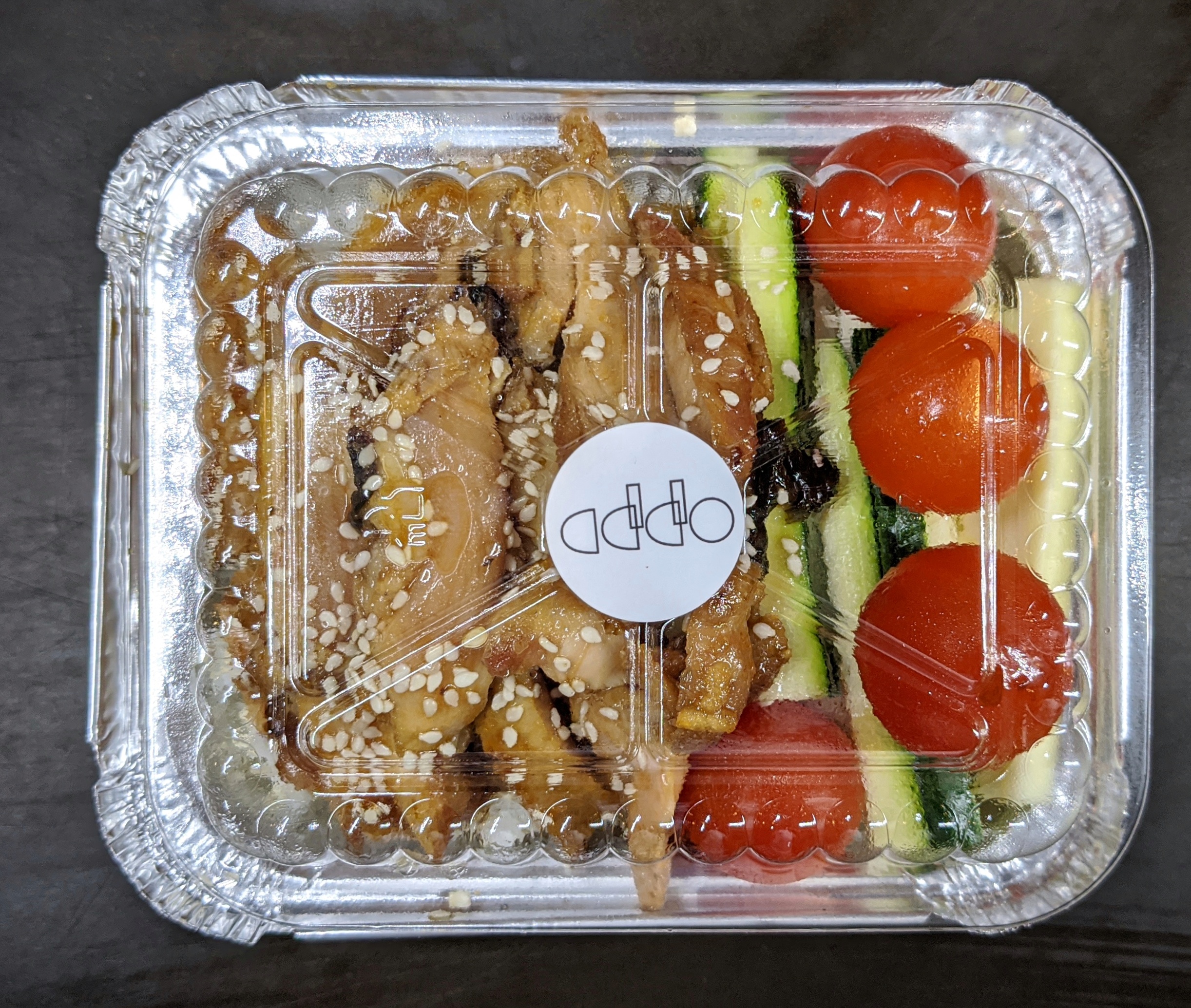 A takeout container with chicken teriyaki, cucumbers, and tomatoes, and a label that says Addo