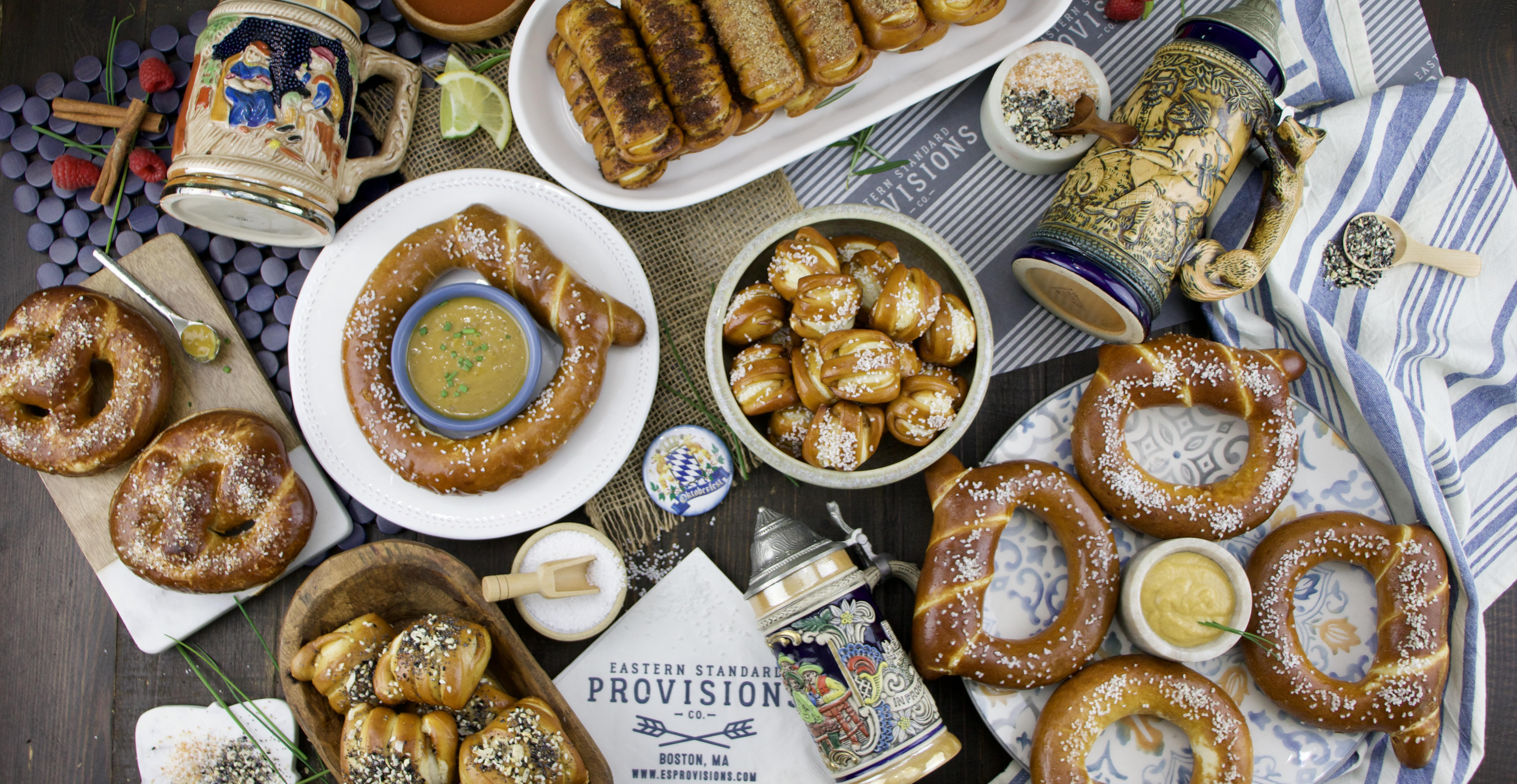 Overhead view of an Oktoberfest-themed selection of several shapes of soft pretzels, dipping sauces, and salts, accompanied by several decorated beer steins