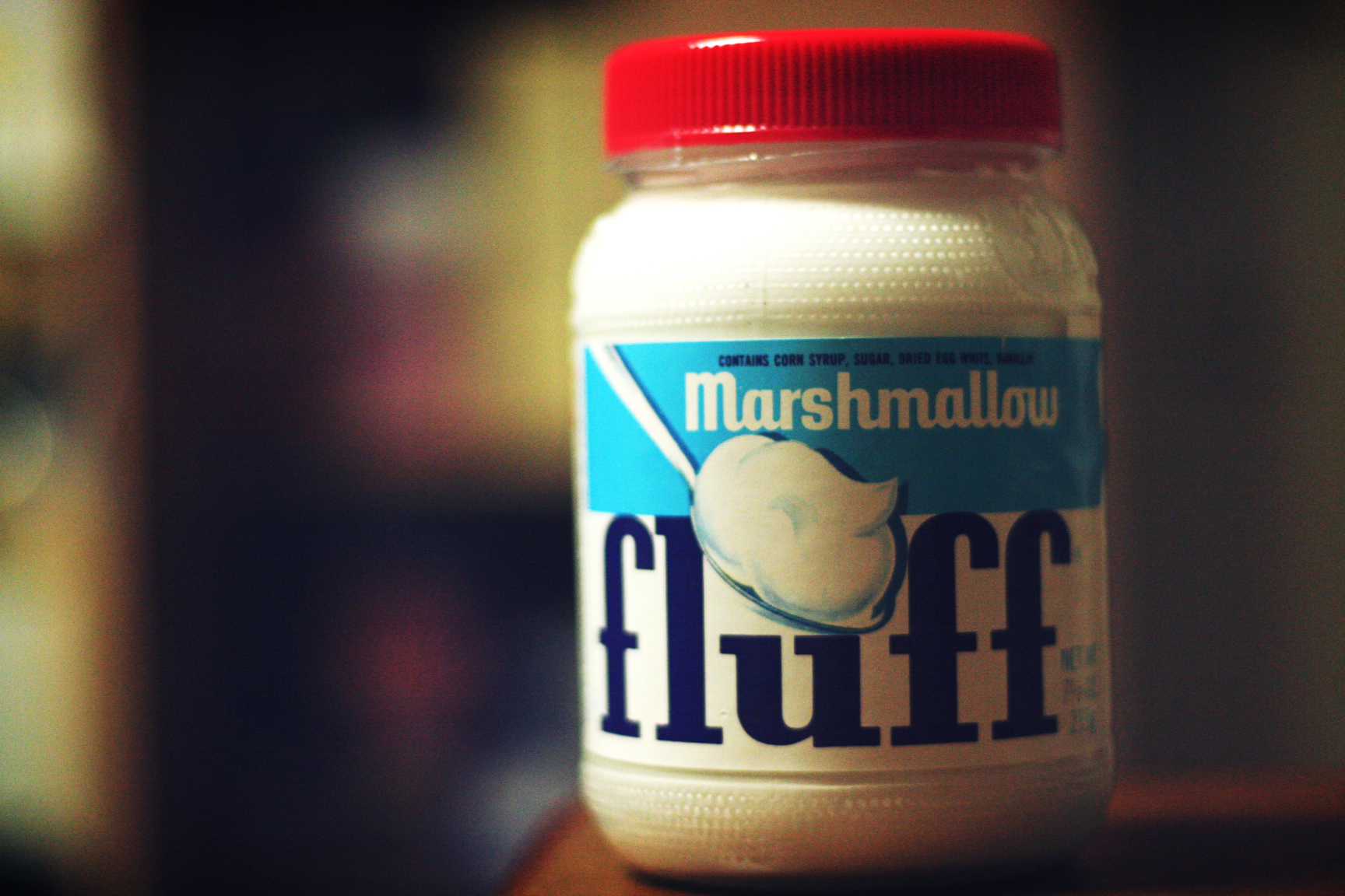 Product shot of a jar of marshmallow Fluff with a red cap and a light blue and white label with dark blue lettering. The jar appears in front of a dark, blurred background.