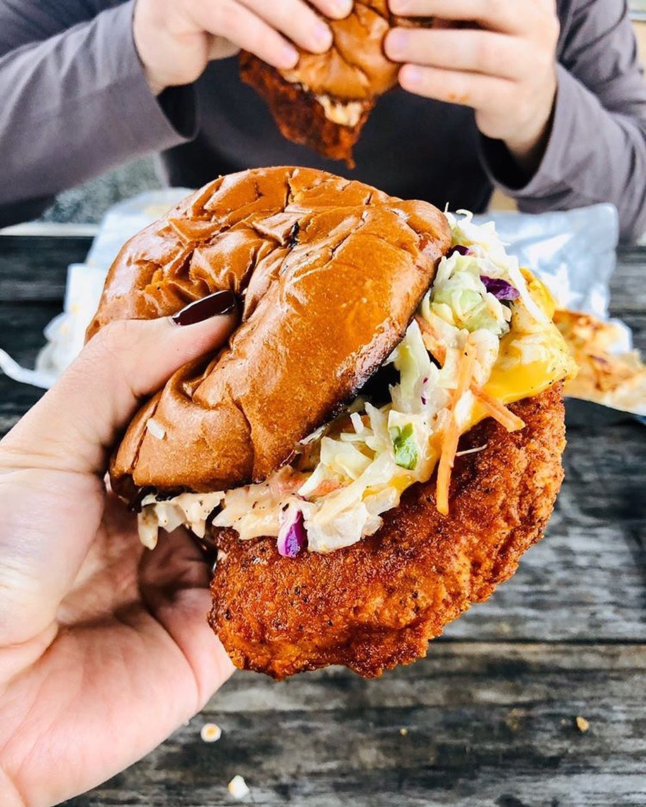 a pale-skinned hand with a dark red manicure holds a messy fried chicken sandwich topped with cole slaw.