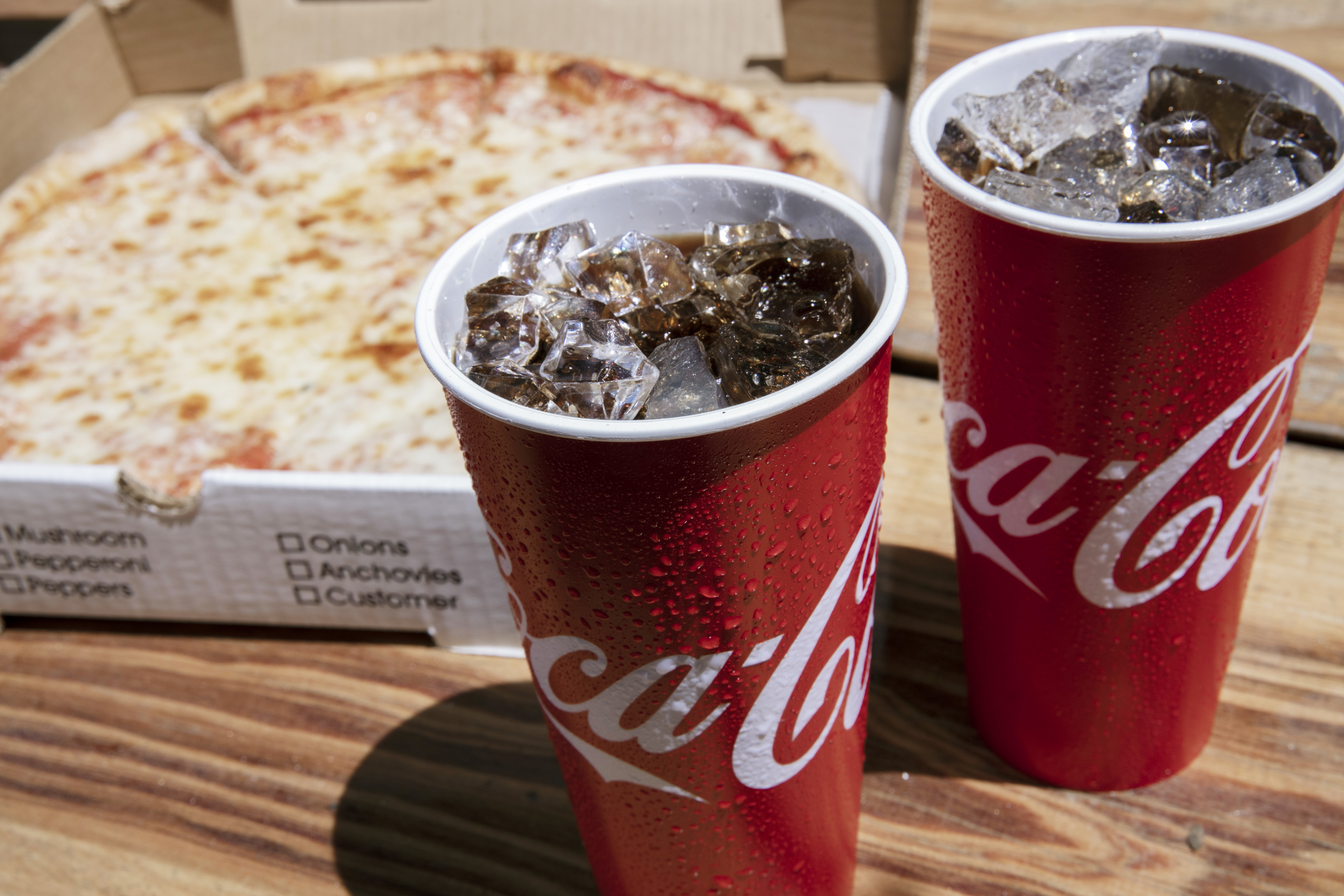 A cheese pizza in a delivery box, plus two paper cups of Coca-Cola.