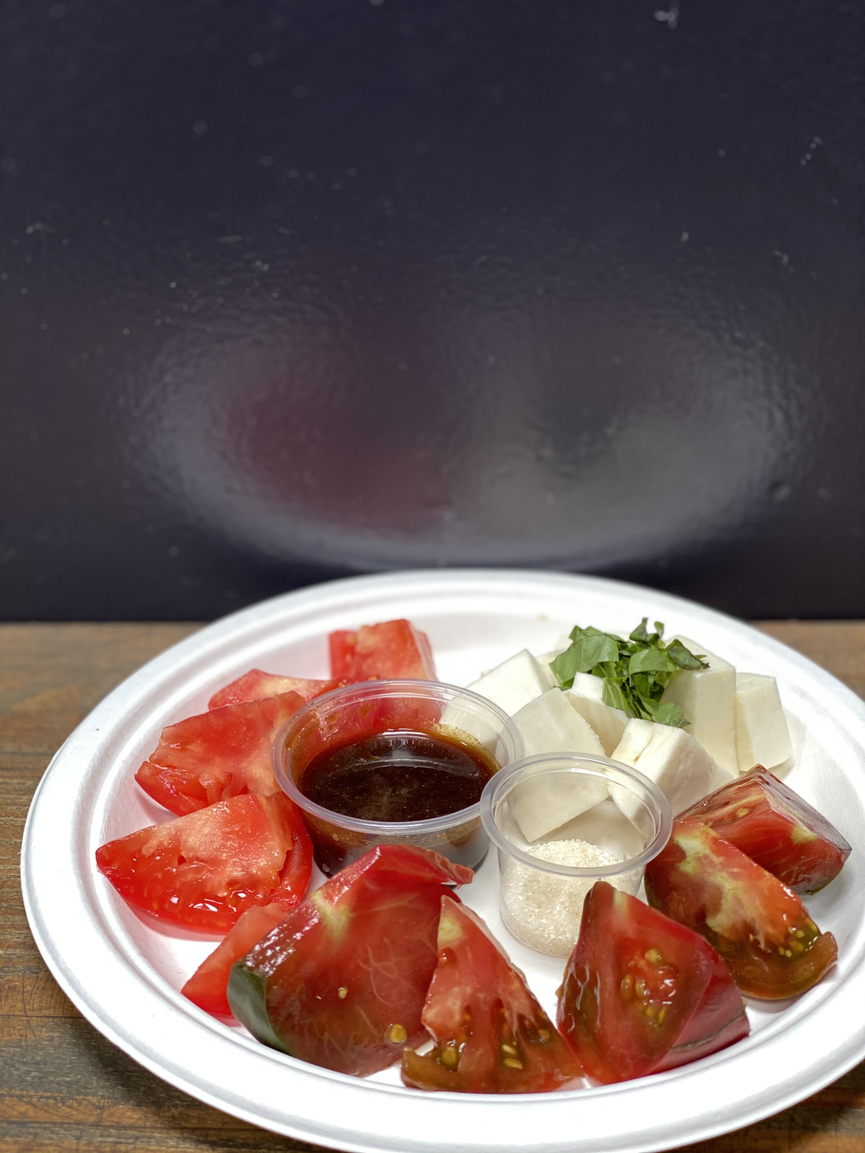 Wedges of juicy red heirloom tomatoes and cubes of mozzarella on a plate, with small tubs of sugar and soy sauce glaze for dipping