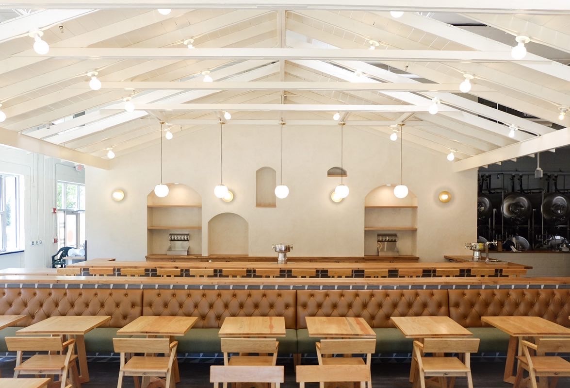 A white vaulted ceiling with exposed beams above the dining room with a row of natural colored two-top tables and chairs and an Army green and camel leather banquet; five pendant lights hang above the bar in the background