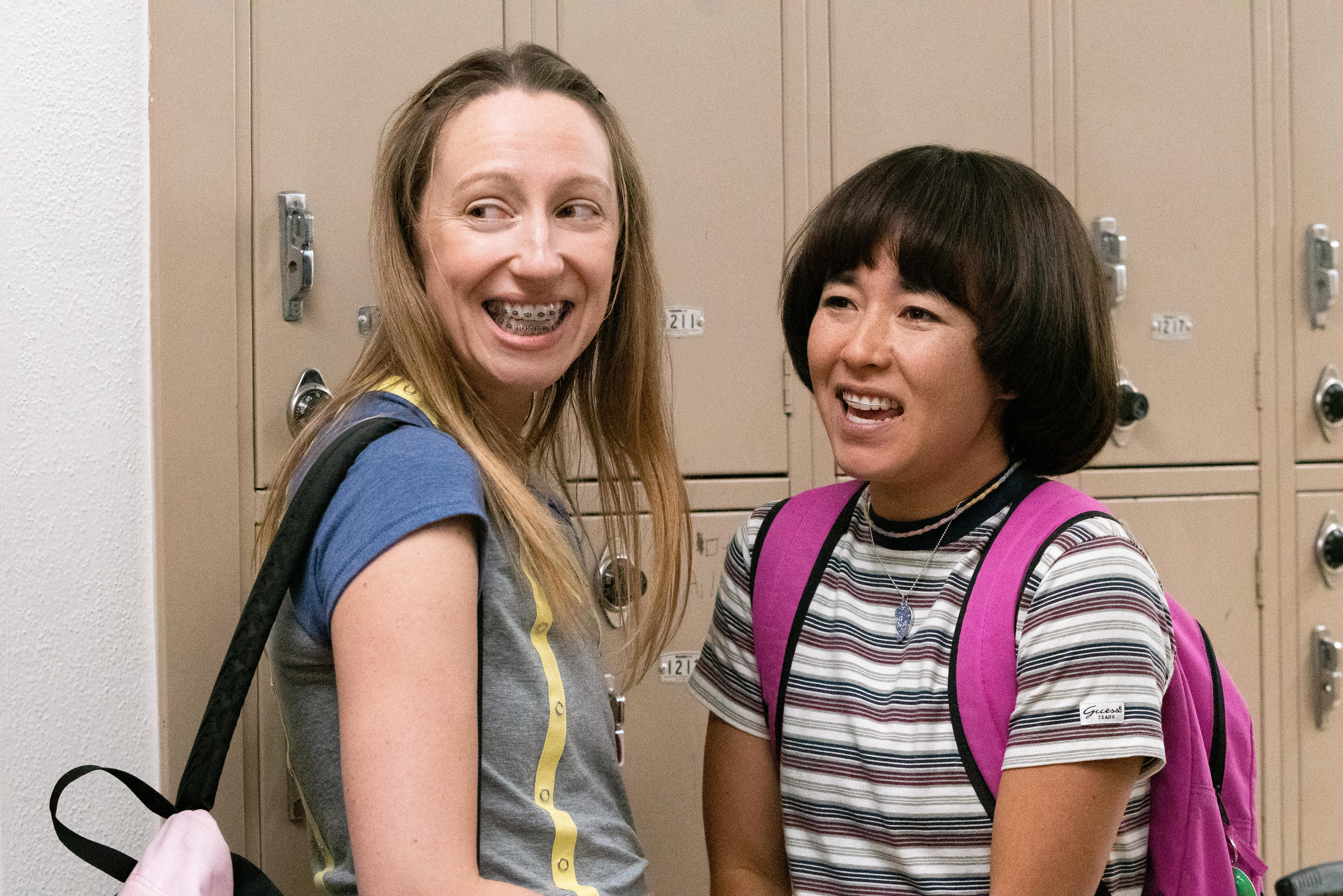 Anna Konkel and Maya Erskine wear backpacks and smile next to a row of tan lockers in Pen15.